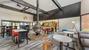 A spacious indoor area with multiple seating arrangements, including tables and chairs, a central fire feature, and a large window providing natural light.