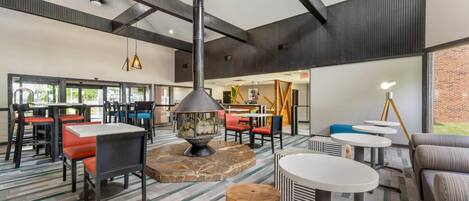 A spacious indoor area with multiple seating arrangements, including tables and chairs, a central fire feature, and a large window providing natural light.