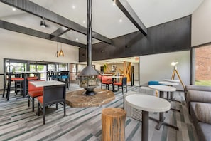 Un vaste espace intérieur aménagé avec différents coins salon, comprenant des tables et des chaises, un foyer central et une grande fenêtre qui laisse entrer la lumière naturelle.