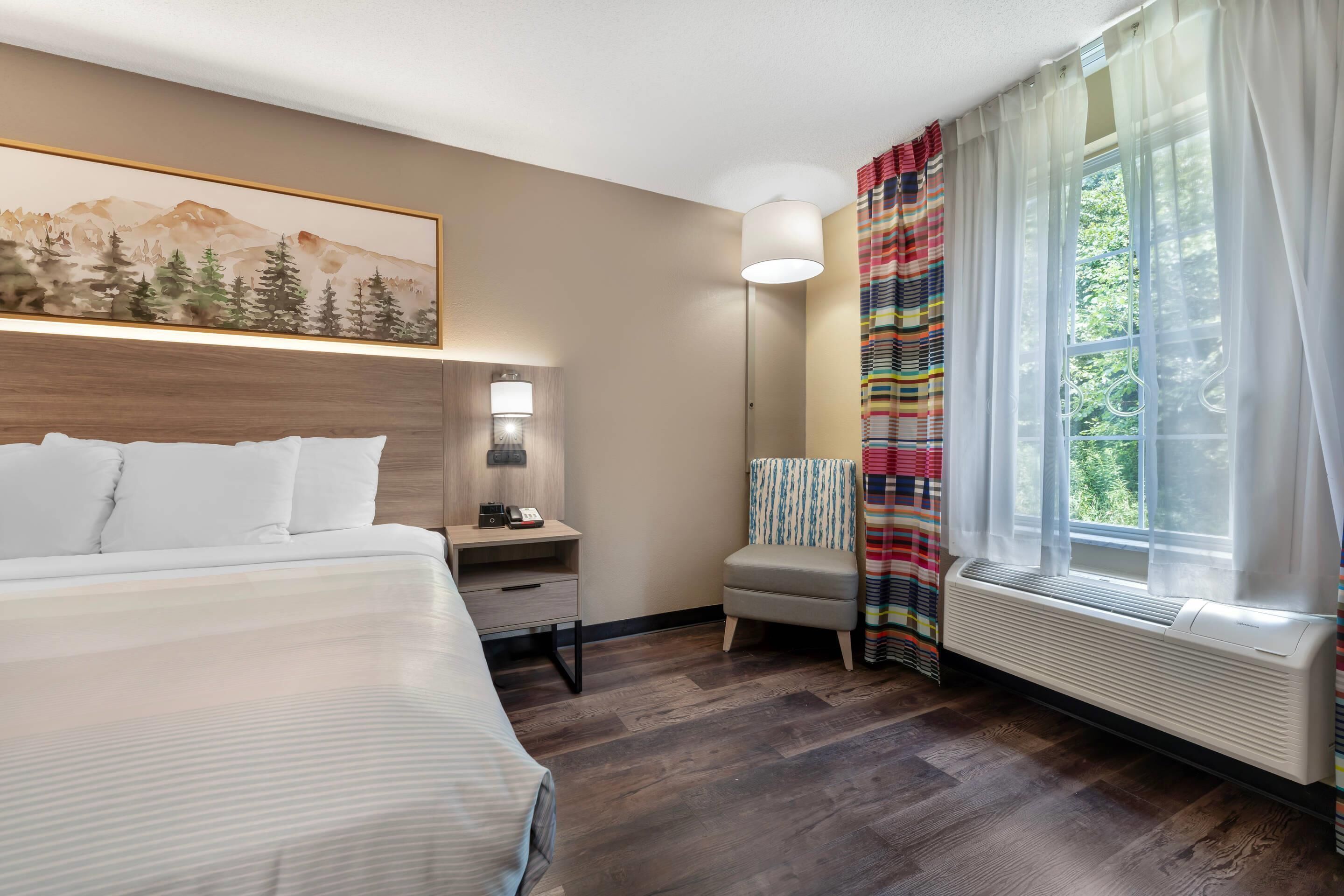 Una habitación de hotel con cama, mesita de noche, lámpara, silla y ventana con cortinas.