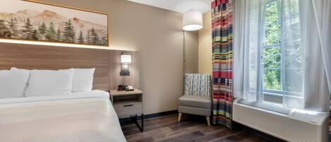 Una habitación de hotel con cama, mesita de noche, lámpara, silla y ventana con cortinas.
