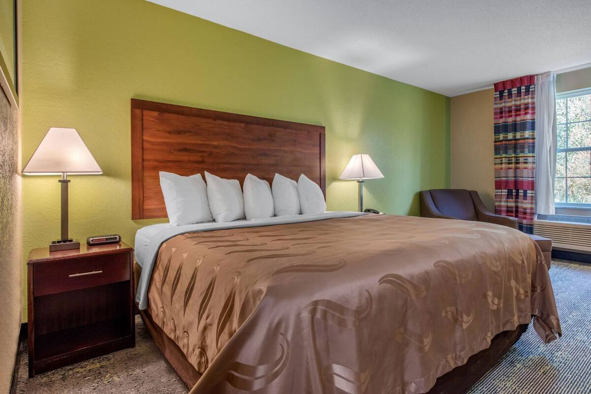 A hotel room with a bed, two bedside lamps, a wooden headboard, a chair, and a window with curtains.