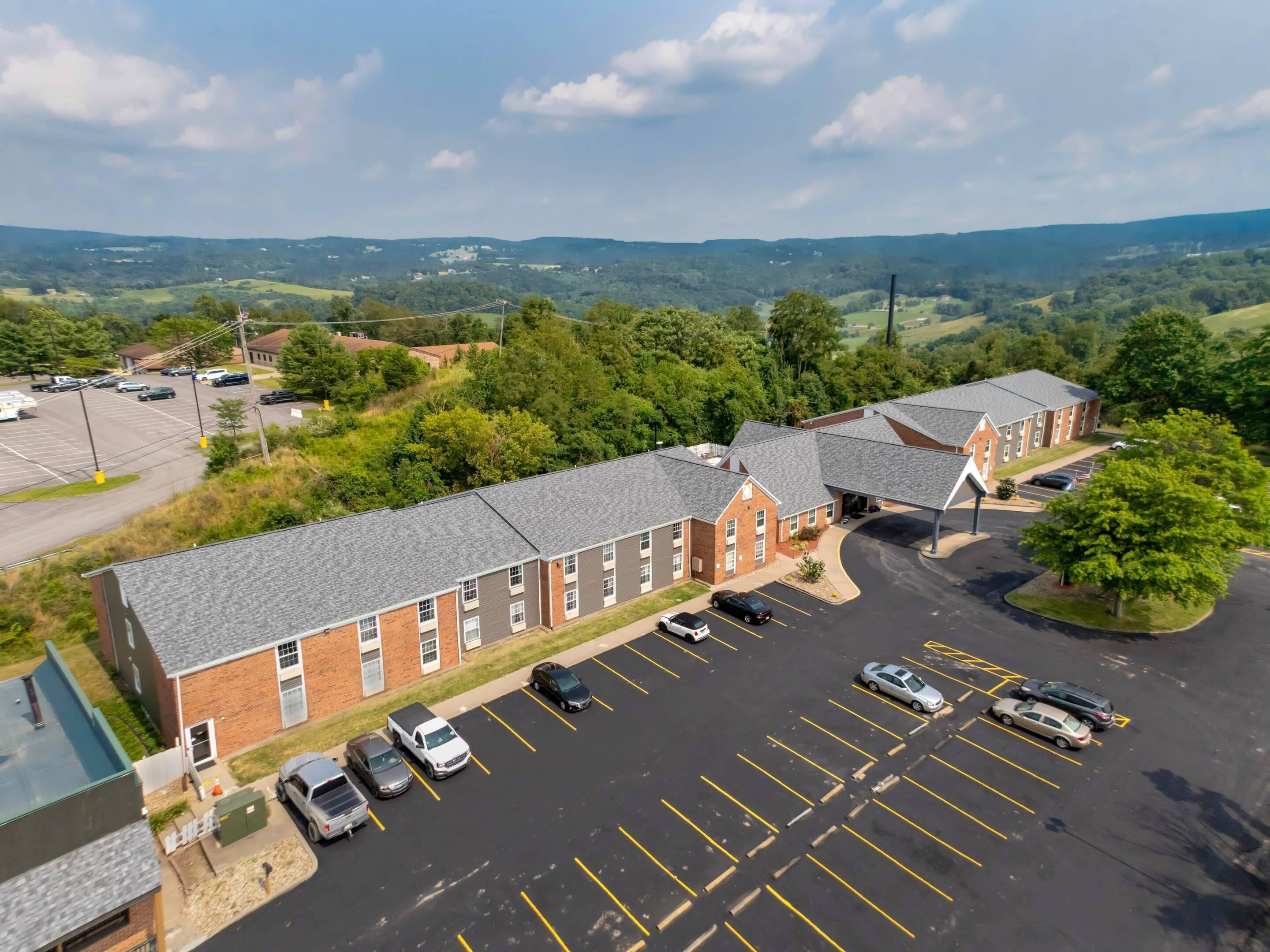 A multi-story building with a parking lot, surrounded by greenery and hills.