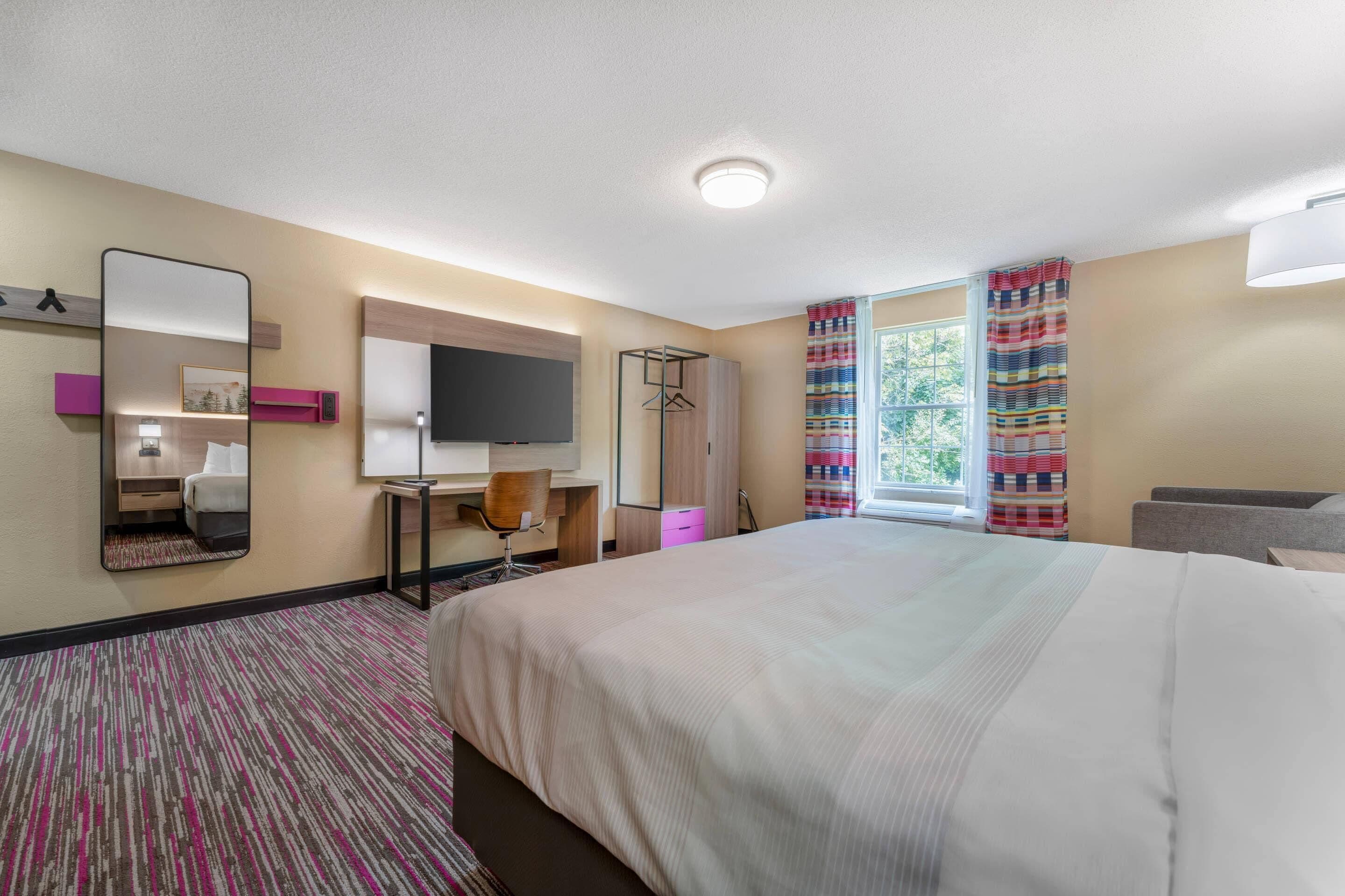 Une chambre d’hôtel équipée d’un lit, d’un bureau, d’une chaise, d’une télévision, d’une armoire et d’une fenêtre avec des rideaux.