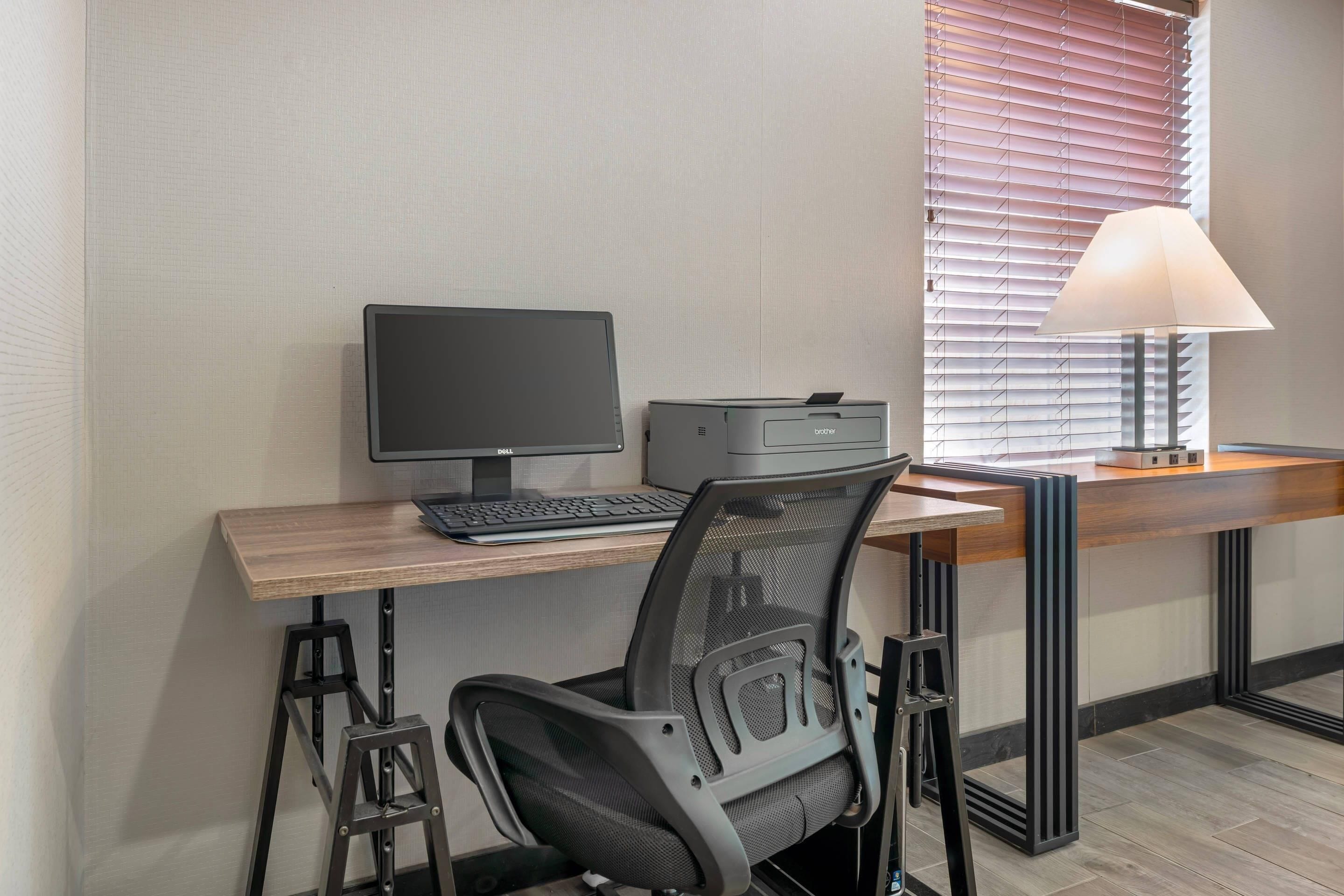 Ein moderner Schreibtisch mit Computer, Drucker und Lampe, platziert in der Nähe eines Fensters mit Jalousien.