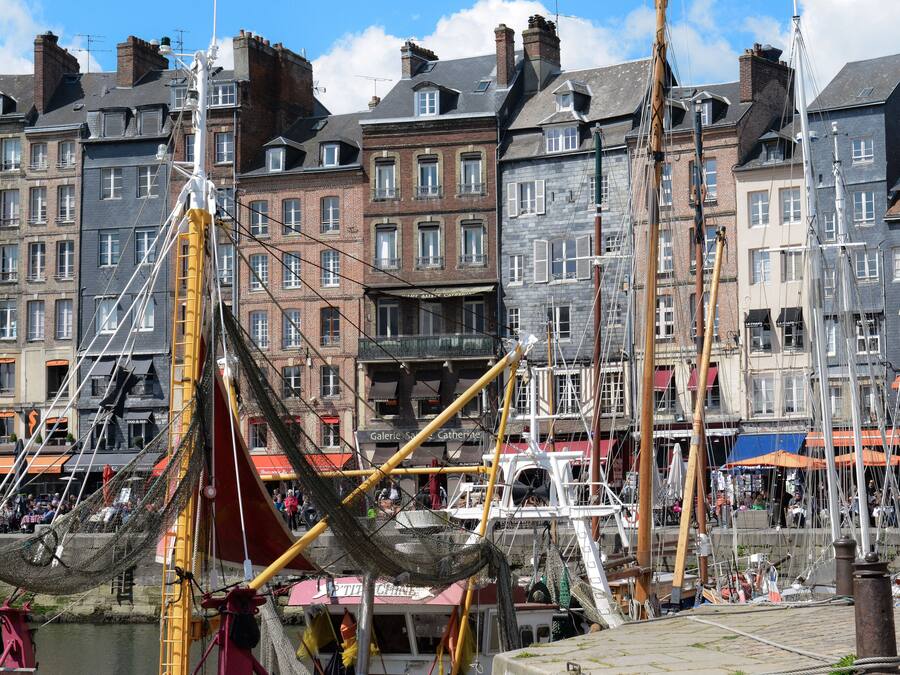 Una zona histórica frente al mar con edificios tradicionales europeos, un barco de vela destacado y una animada escena portuaria.