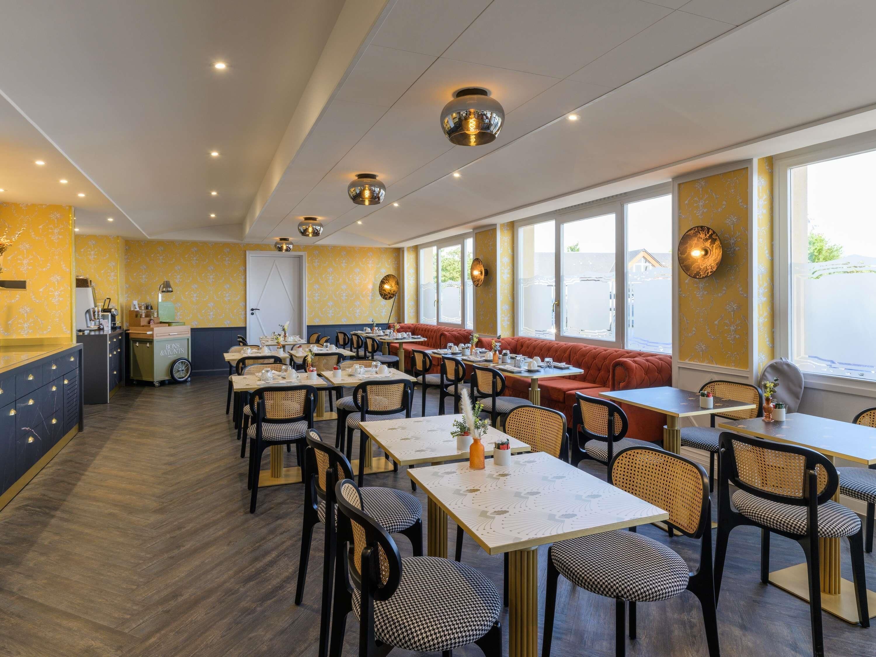 A restaurant with multiple tables and chairs, a kitchen area, and large windows.