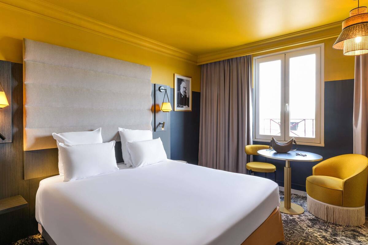 A hotel room with a large bed, a small round table, a chair, and a window with curtains.