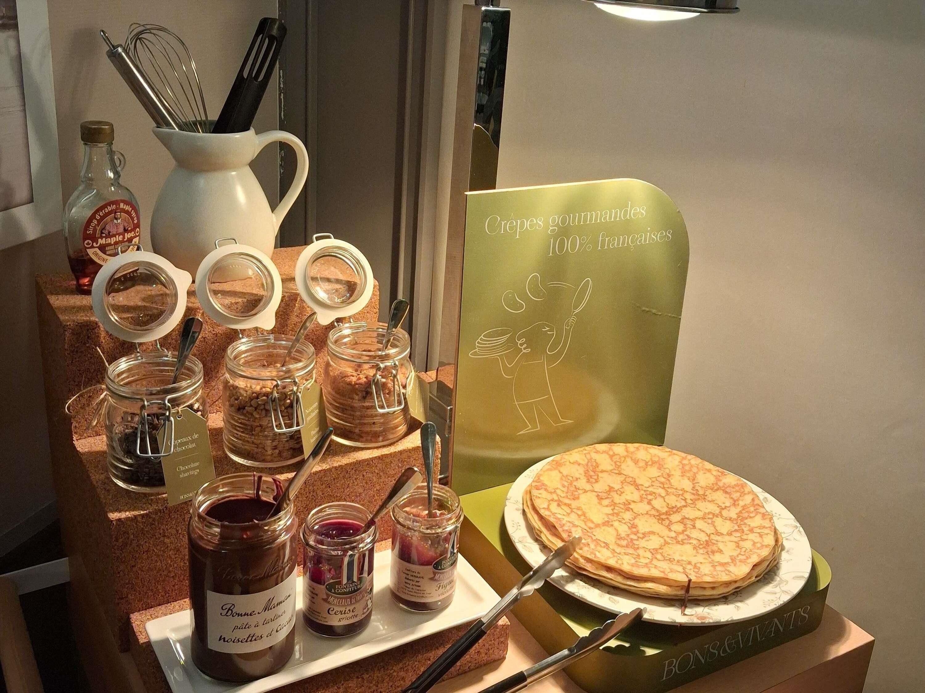 A display of food items including jars of chocolate and fruit preserves, a plate of pancakes, and a sign indicating gourmet French crepes.