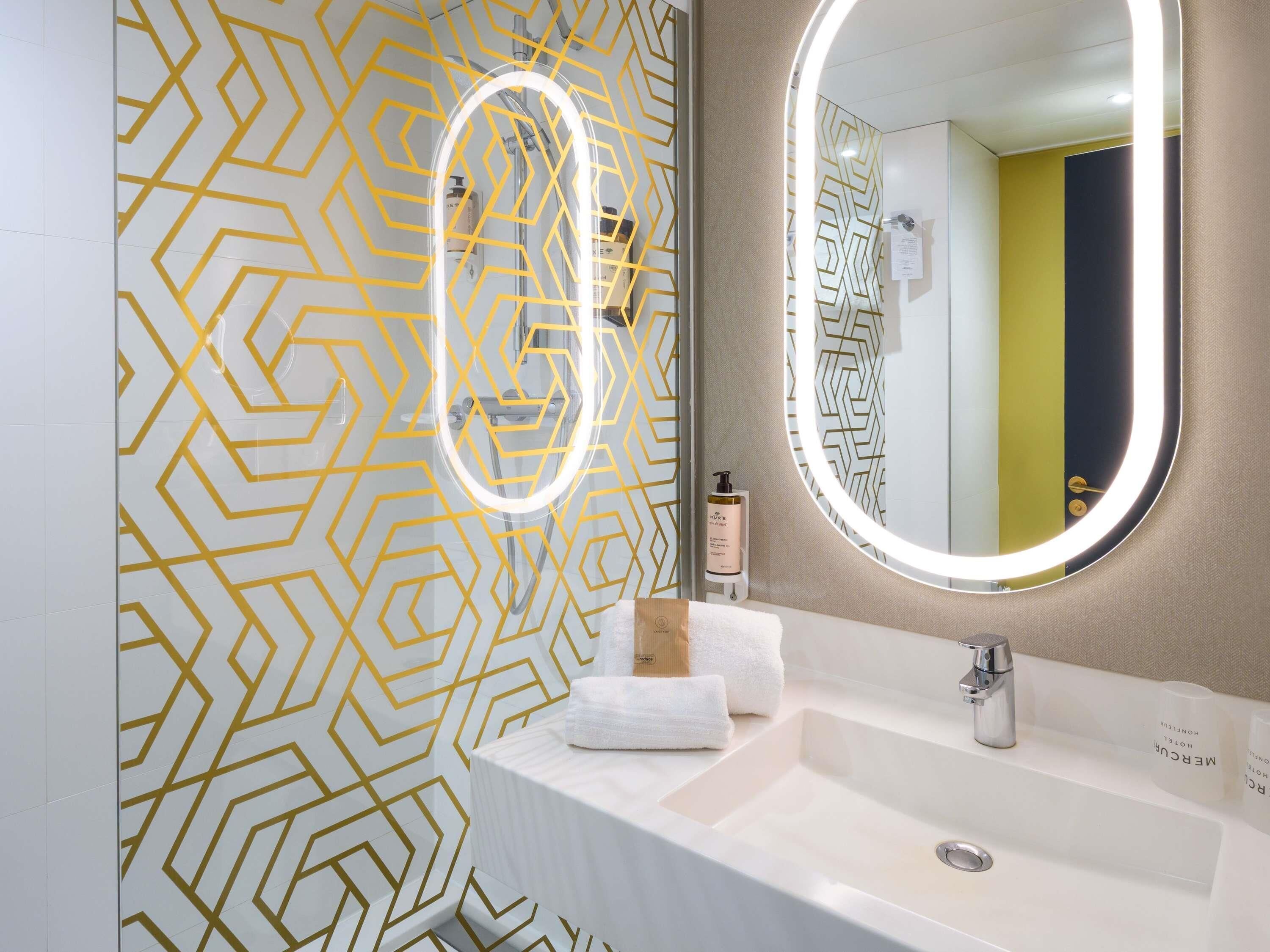 Un baño moderno con pared de diseño geométrico, un espejo redondo con luz y un lavabo blanco.