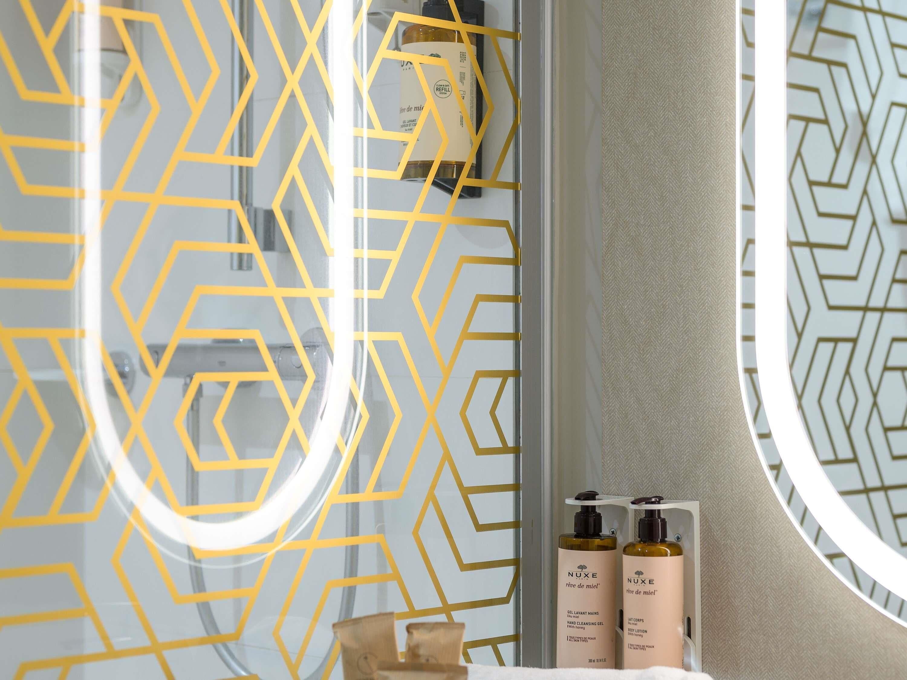 Un baño con pared de diseño geométrico, un espejo redondo y dos botellas de crema corporal Nuxe en un estante.