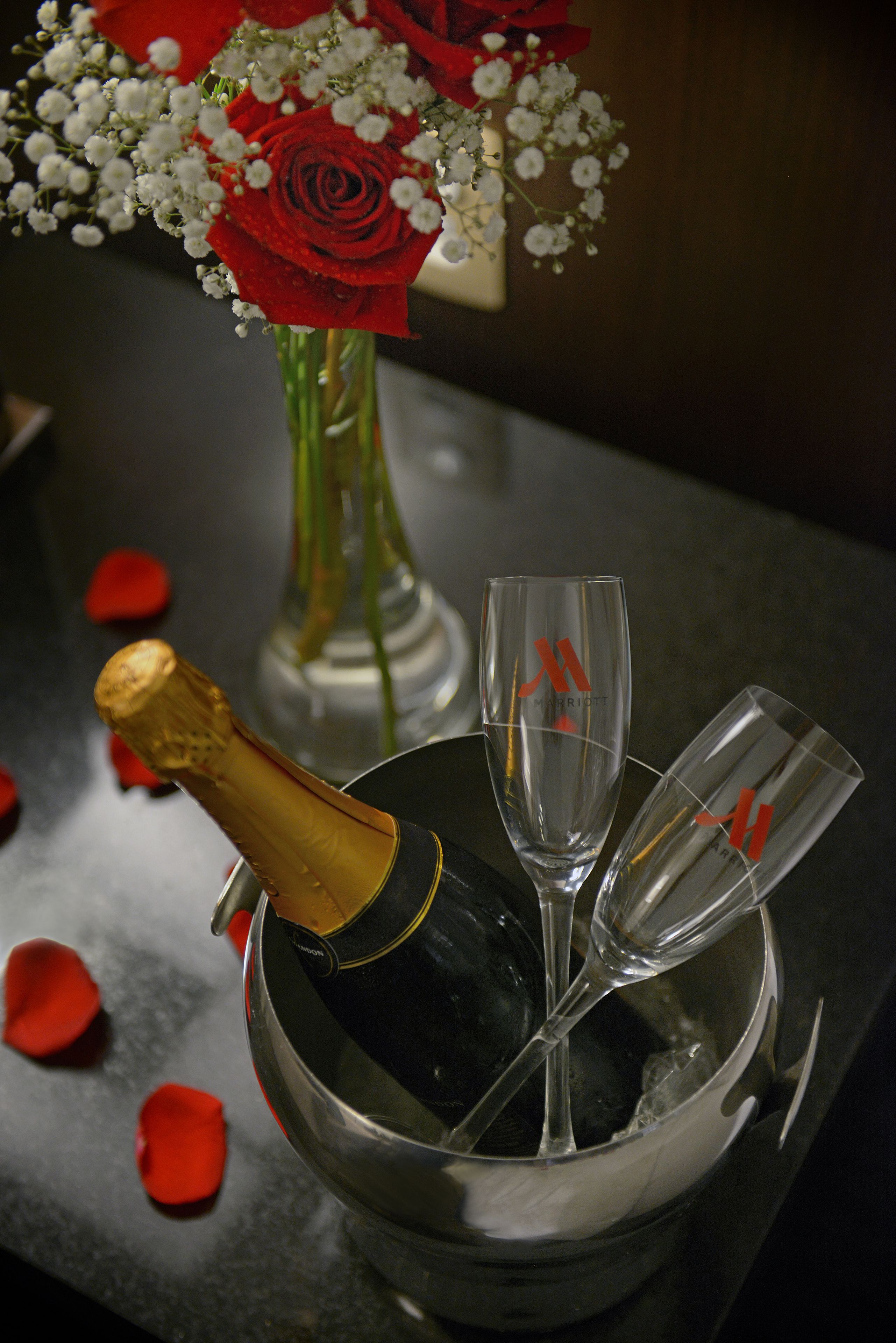 A bottle of champagne in a bucket with two glasses and a bouquet of red roses.