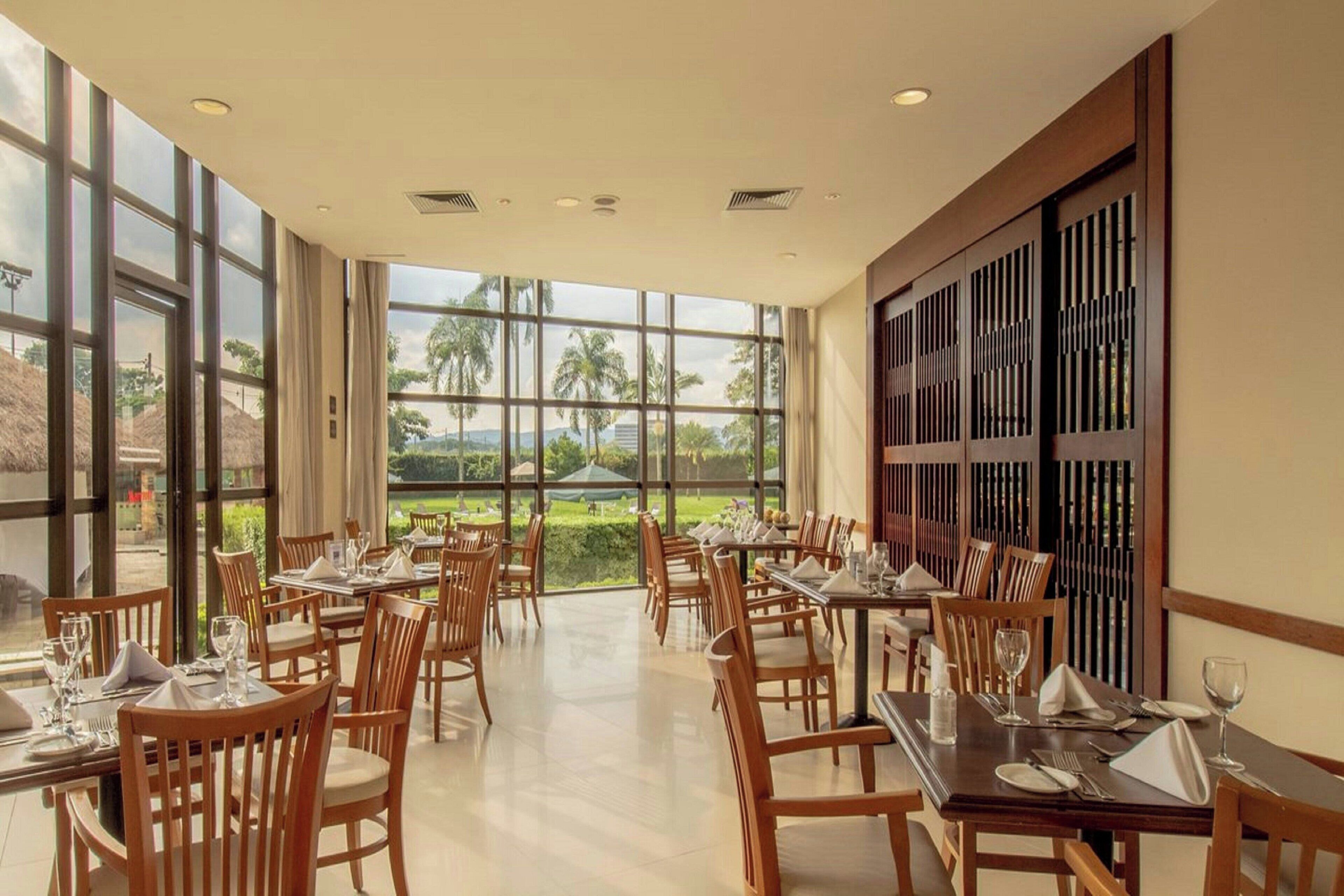 Um restaurante com várias mesas e cadeiras, grandes janelas que oferecem vista para palmeiras e vegetação, e uma divisória de parede de madeira tradicional.