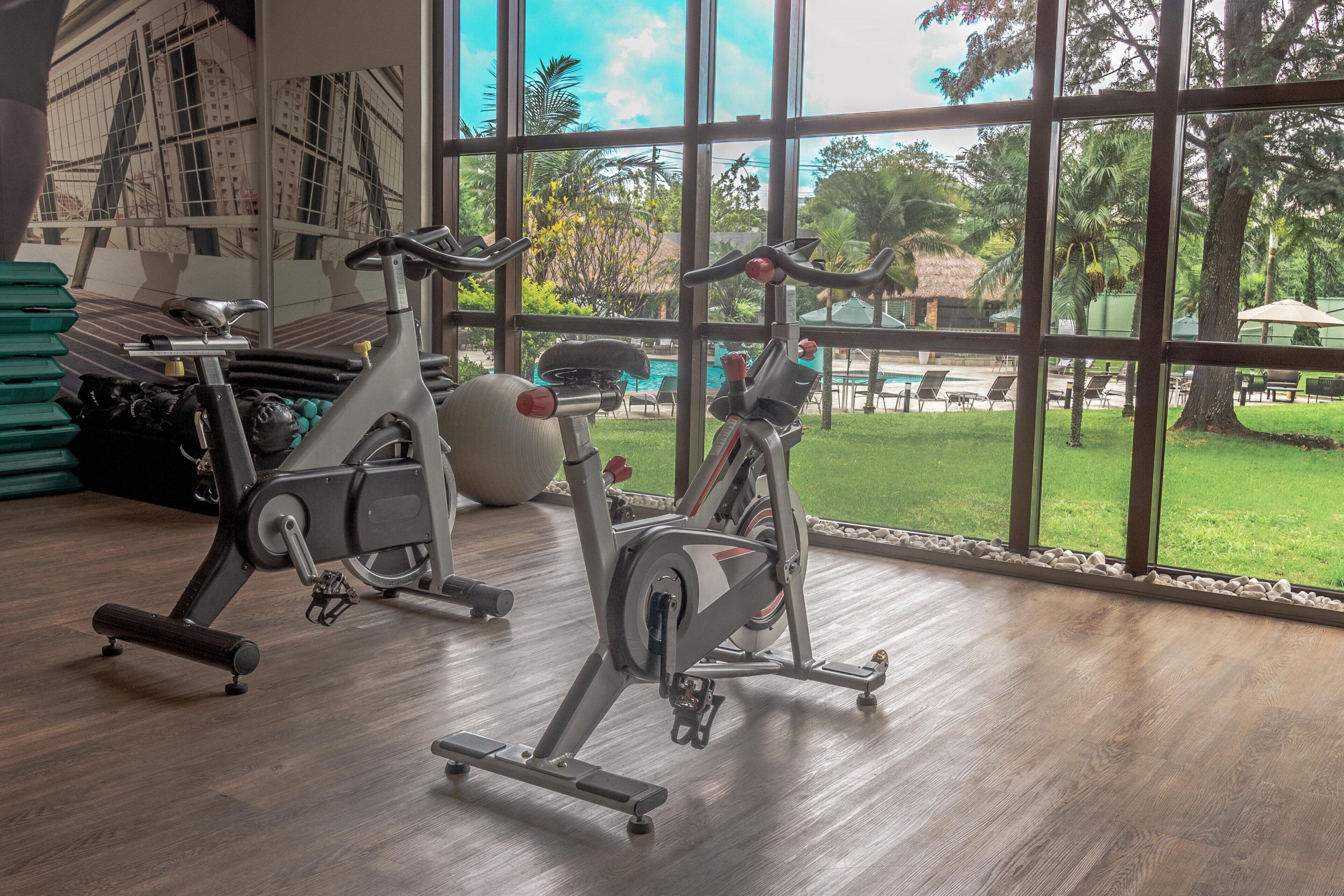 A gym with stationary bikes and exercise balls, large windows with a view of a pool and greenery.