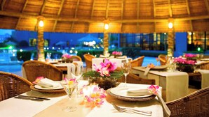 A restaurant with tables set for dining, featuring wicker chairs and a thatched roof.