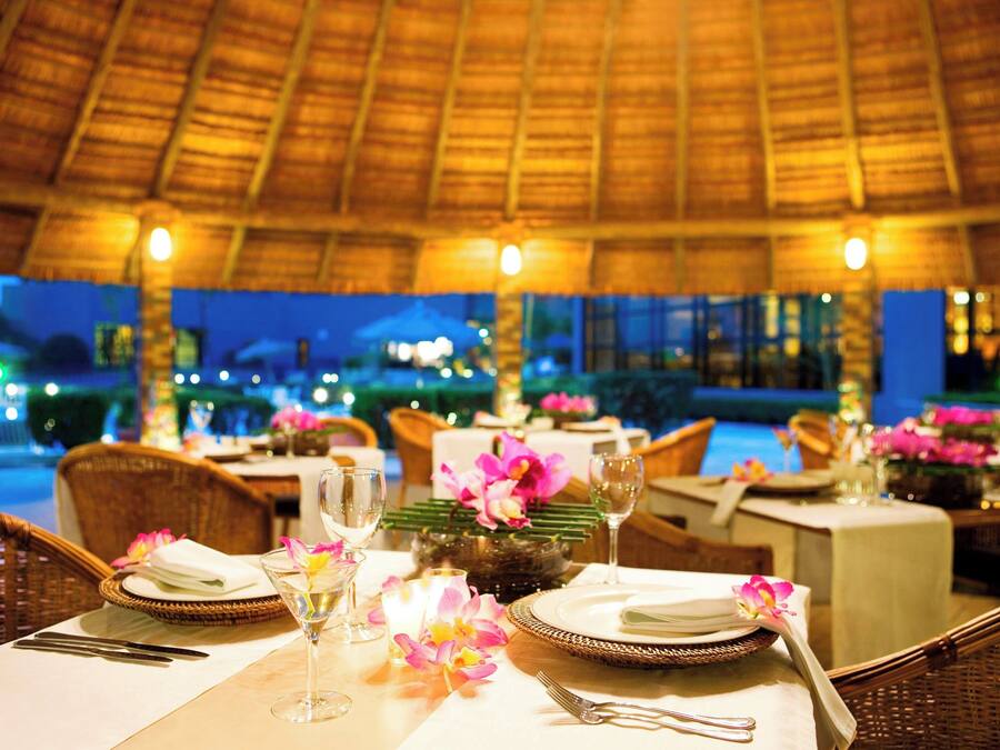 A restaurant with tables set for dining, featuring wicker chairs and a thatched roof.