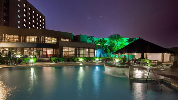 A nighttime view of a hotel with a swimming pool, outdoor seating, and a building with multiple floors and illuminated windows.