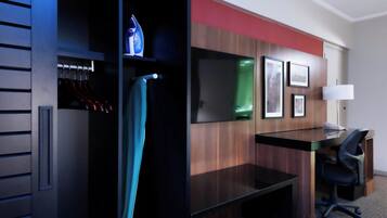 Quarto de hotel com escrivaninha de madeira, uma cadeira, guarda-roupa com roupas penduradas e uma cama.