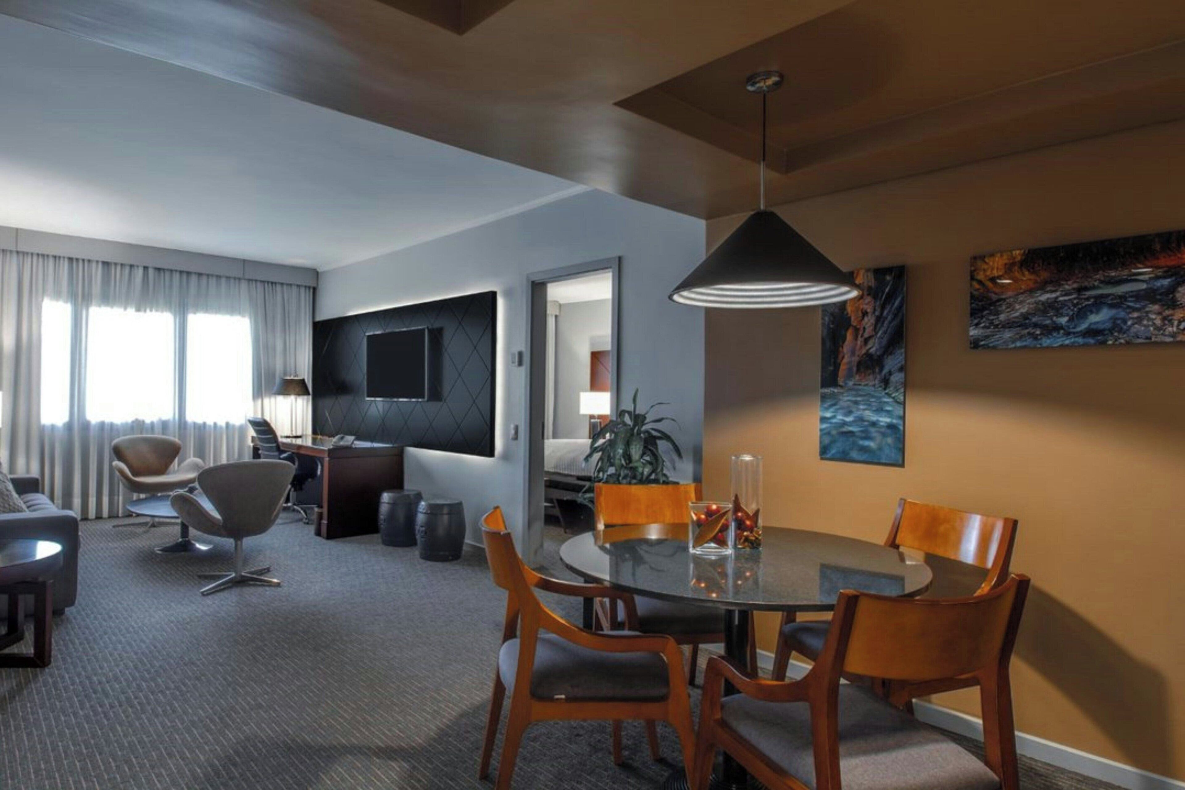 Una habitación de hotel moderna con zona de comedor, televisor de pantalla plana, planta y obras de arte en las paredes.