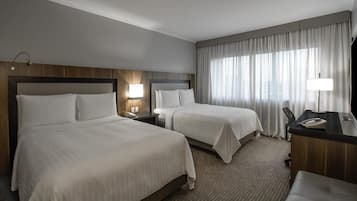 A hotel room with two beds, a desk, a chair, a lamp, and a window with curtains.