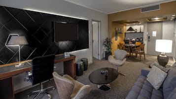 Una habitación de hotel moderna con una televisión de pantalla plana montada en una pared negra, un escritorio con una lámpara, un sofá, una mesa de centro y una zona de comedor con sillas y una mesa.
