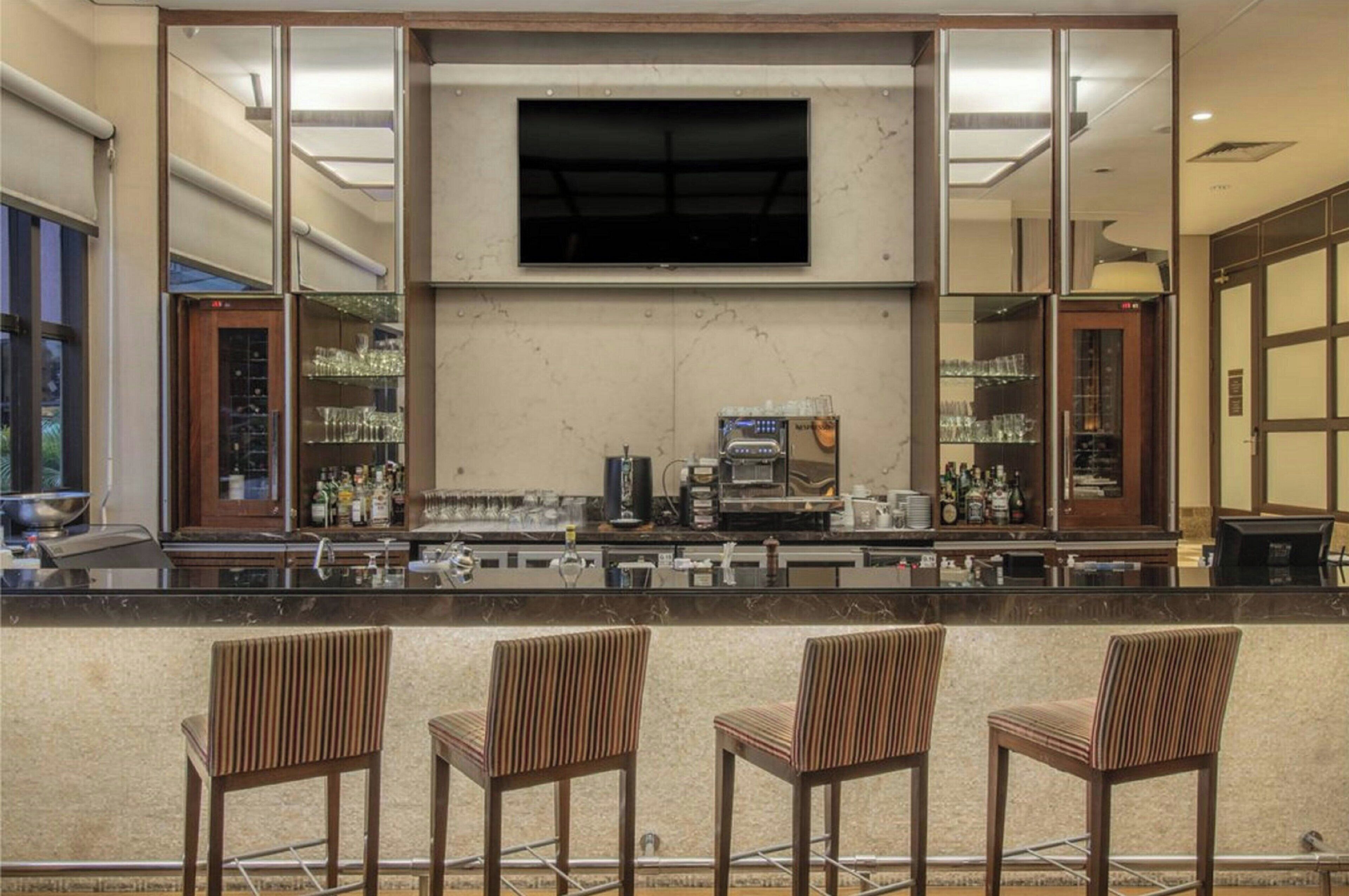 A bar area with a flat-screen TV mounted above the counter, a coffee machine, and shelves with glassware and bottles. There are three striped bar stools in front of the counter.