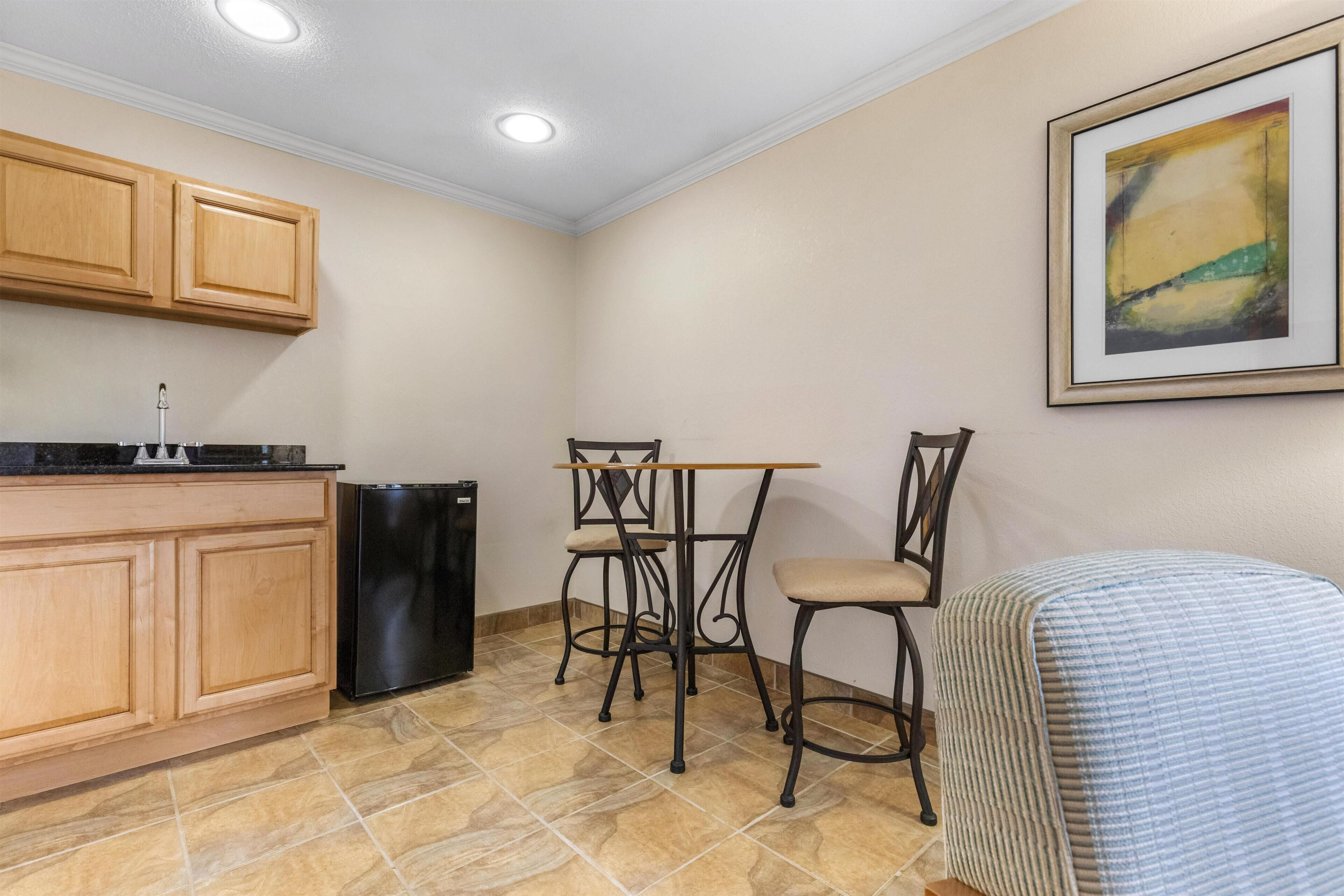 A compact kitchen with wooden cabinets, a black refrigerator, a dining table with chairs, and a framed abstract painting on the wall.
