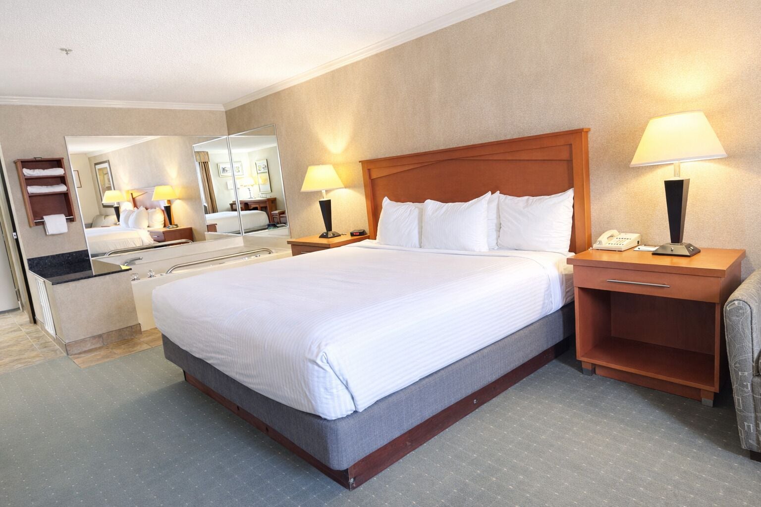A hotel room with a large bed, two bedside tables with lamps, a mirror, a bathroom with a sink and mirror, and a small seating area with a chair.