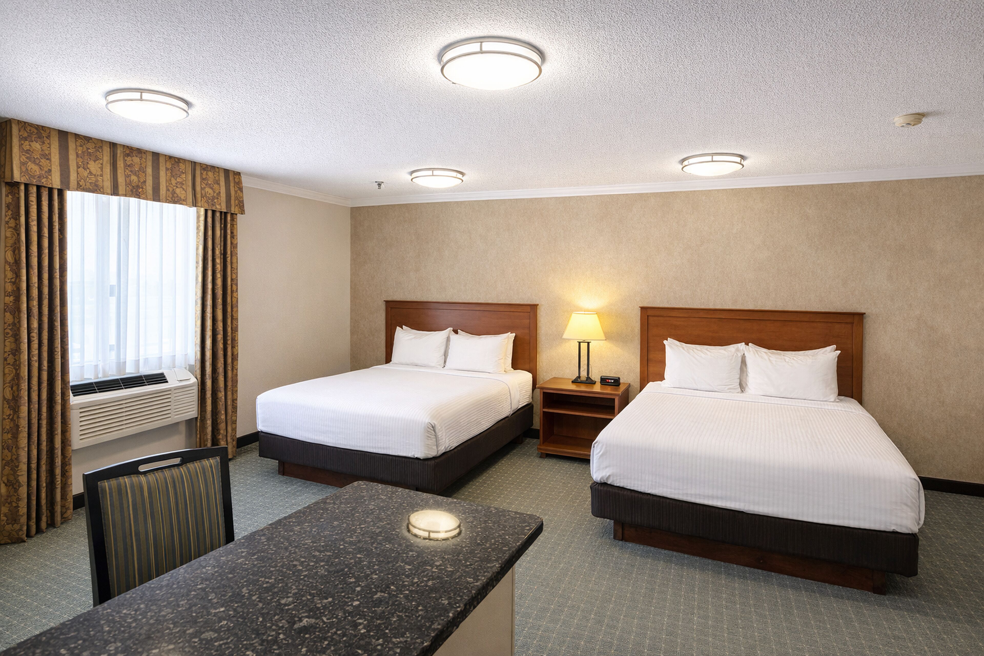 Une chambre d’hôtel avec deux lits, un bureau, une chaise, une fenêtre avec des rideaux et un plafond doté d’un éclairage encastré.