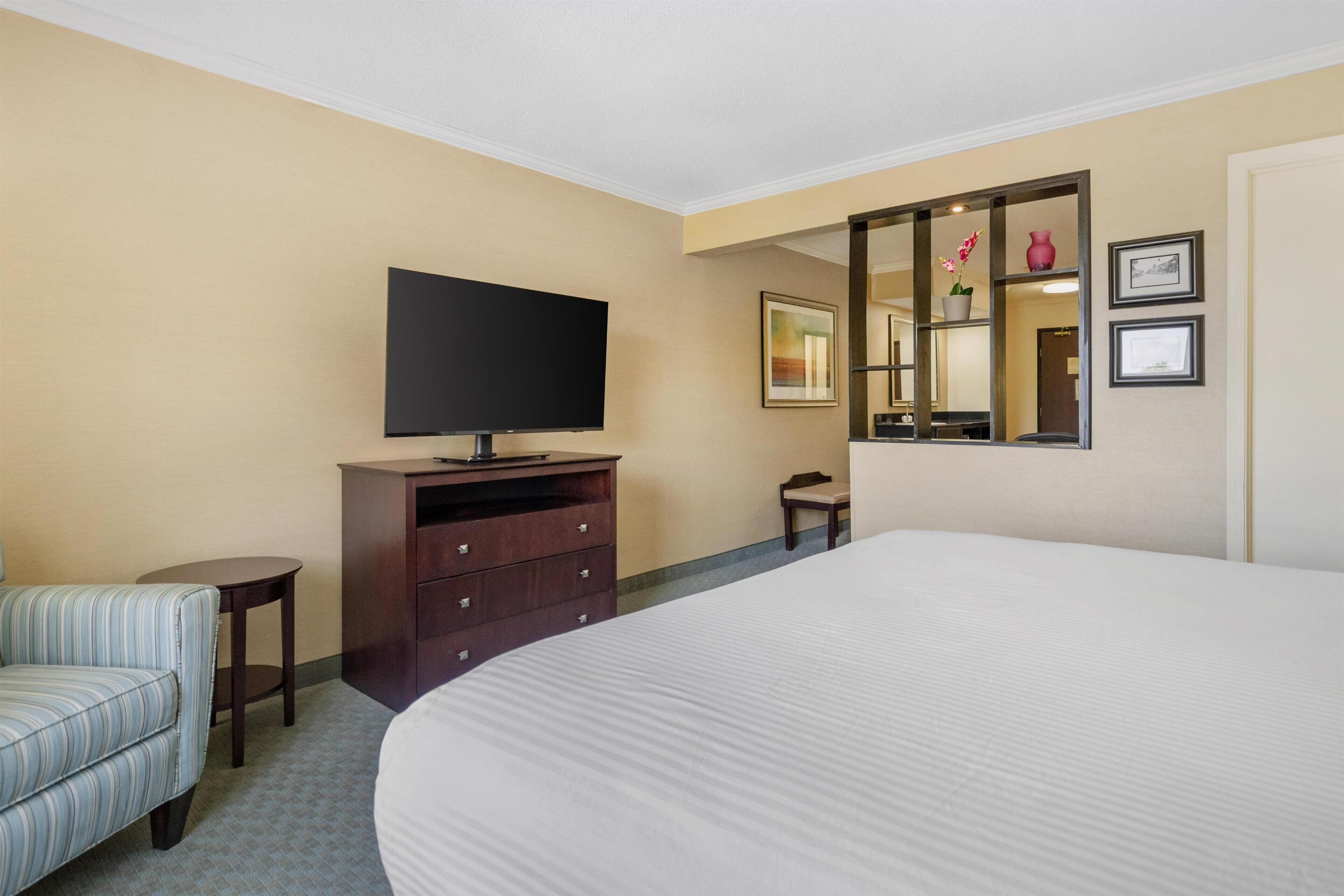 A hotel room with a large bed, a television on a wooden dresser, a small round table, a striped chair, a mirror, and framed pictures on the wall.