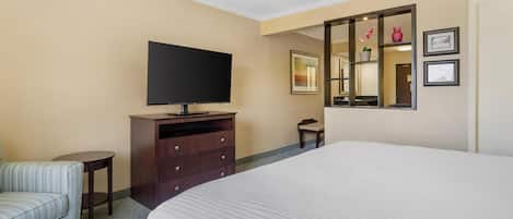 A hotel room with a large bed, a television on a wooden dresser, a small round table, a striped chair, a mirror, and framed pictures on the wall.