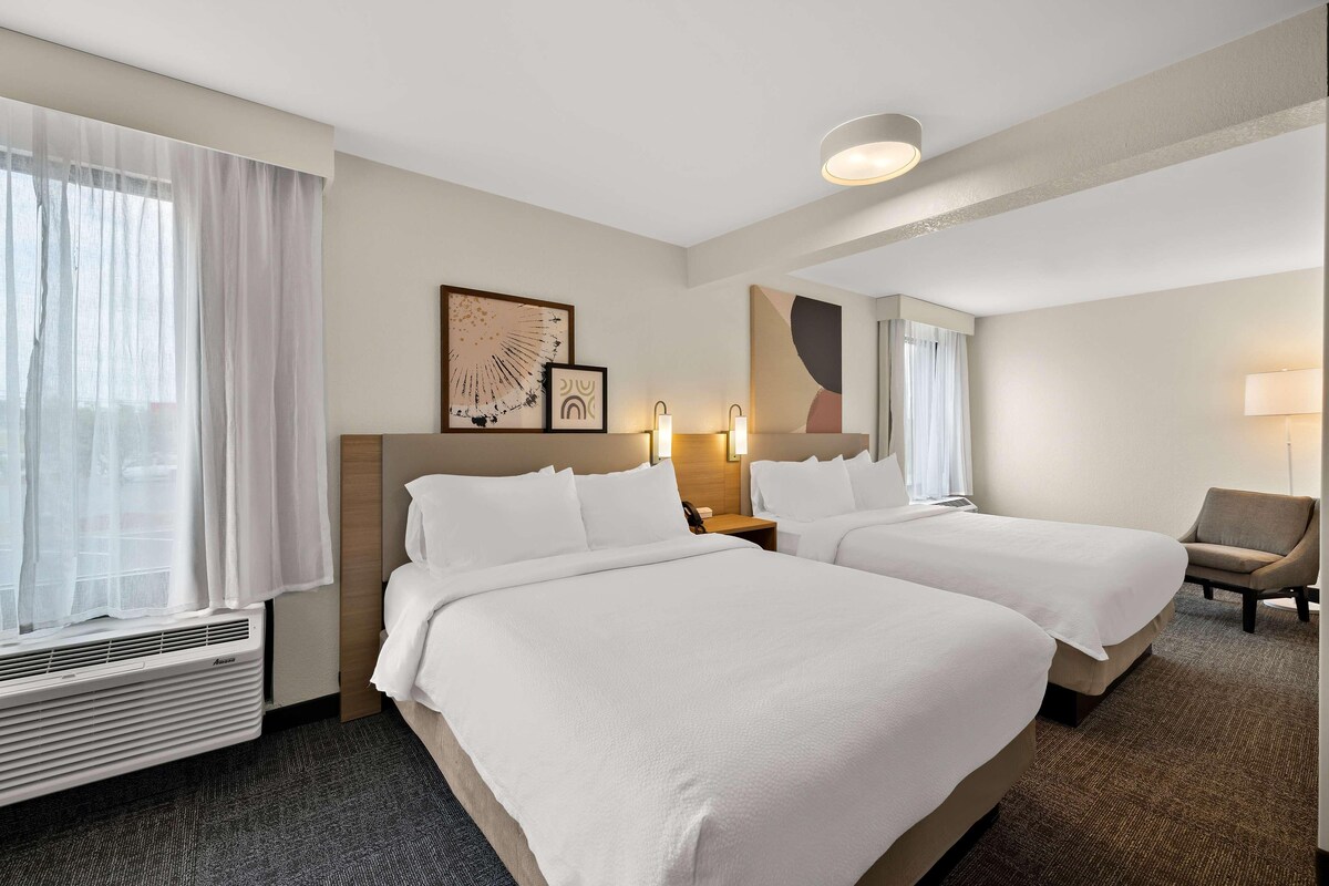 A hotel room with two beds, a chair, a desk, a mirror, and artwork on the wall.