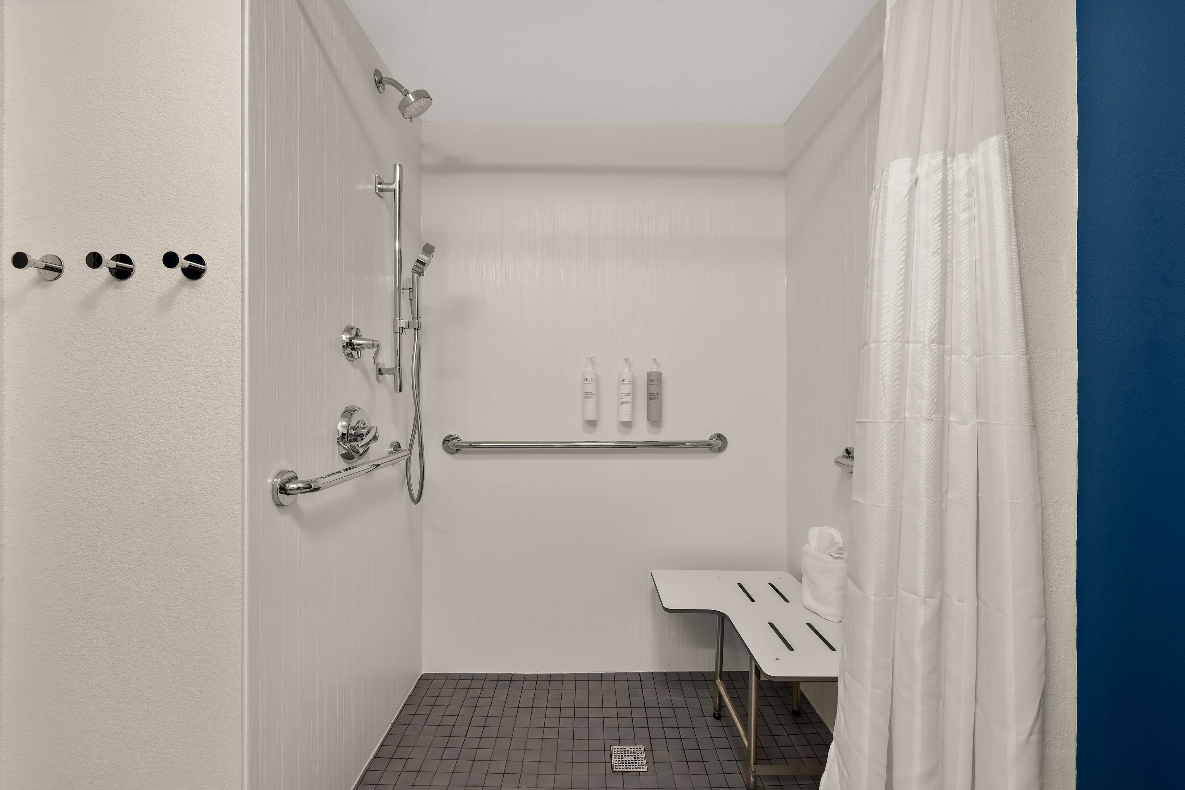 A small, modern bathroom with a shower, a handrail, a towel rack, and a shelf with toiletries.
