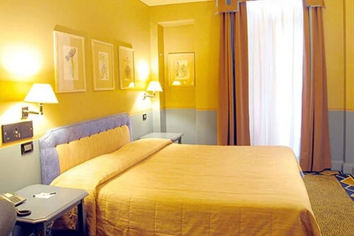 Hotellihuone, jossa on sänky, yöpöydät, lamput ja seinällä tauluja.