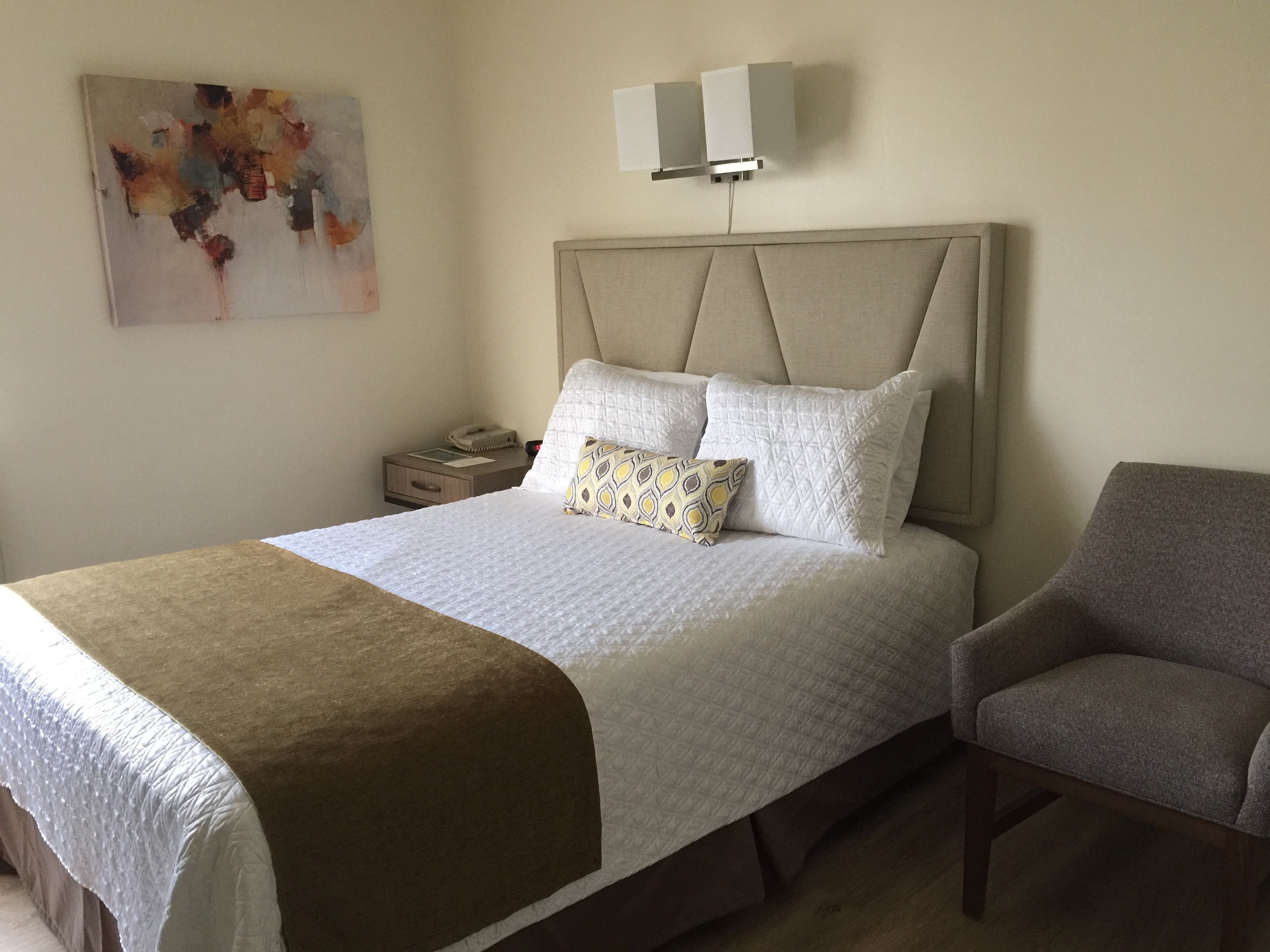 Una cama bien hecha con cabecera, una mesita de noche con lámpara y una silla.
