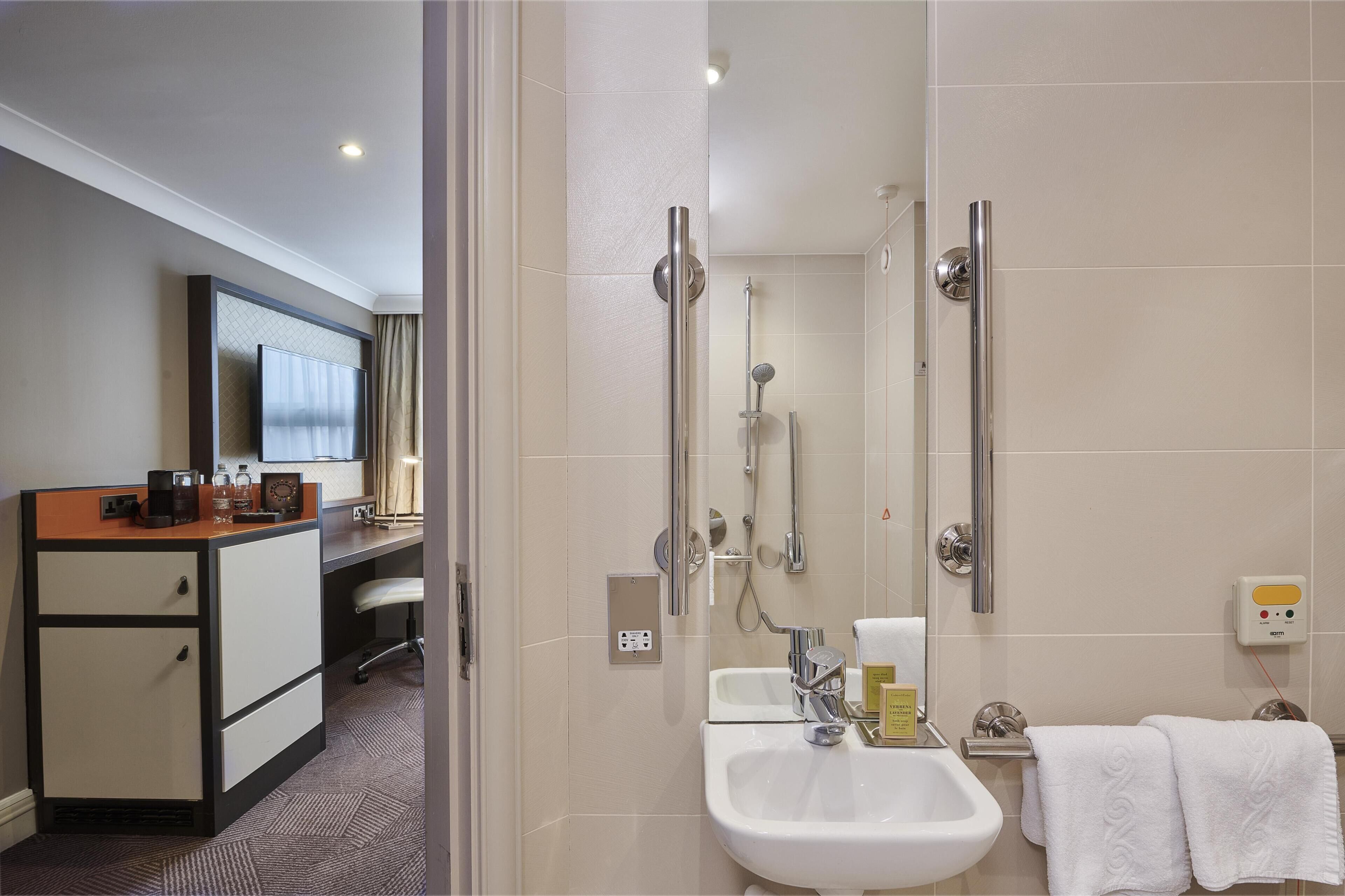 A hotel bathroom with a shower, sink, and a vanity with a mirror.