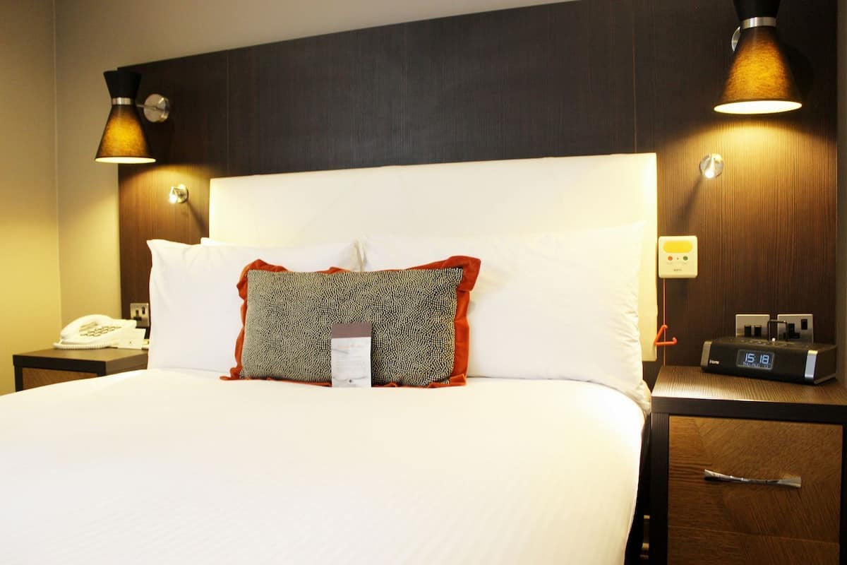 A hotel room with a large bed, two bedside tables, a nightstand with a phone, a digital clock, and two wall-mounted lamps.
