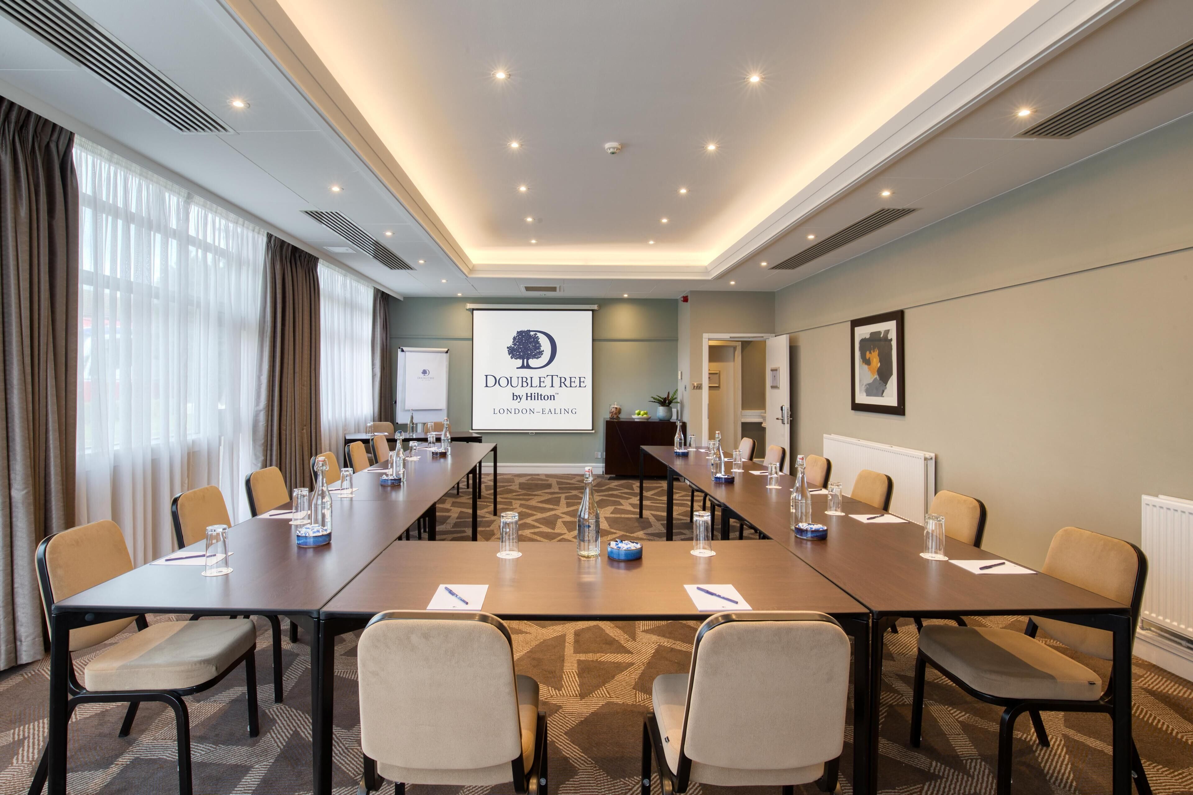 A conference room with a long table, chairs, and a projector screen displaying 'DOUBLETREE by Hilton London-Ealing'.