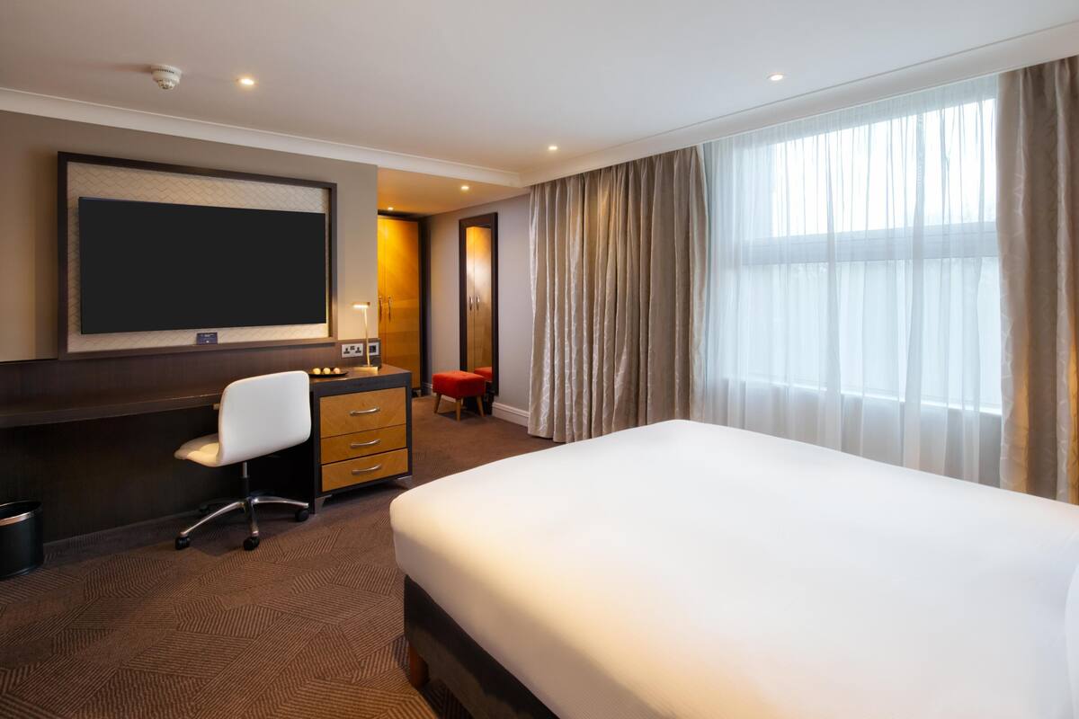 A hotel room with a large bed, a desk with a chair, a wardrobe, and a television.