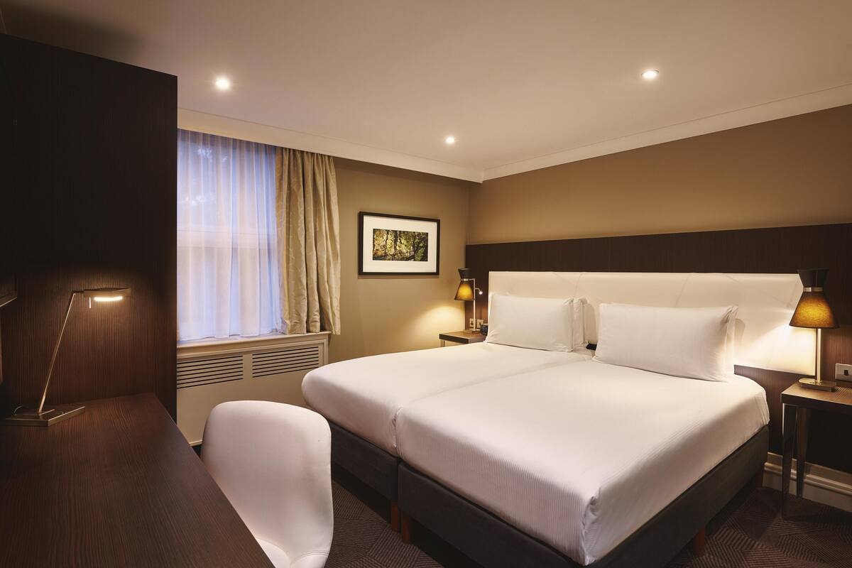 A hotel room with a large bed, a desk, a chair, a window with curtains, a framed picture on the wall, and two bedside lamps.