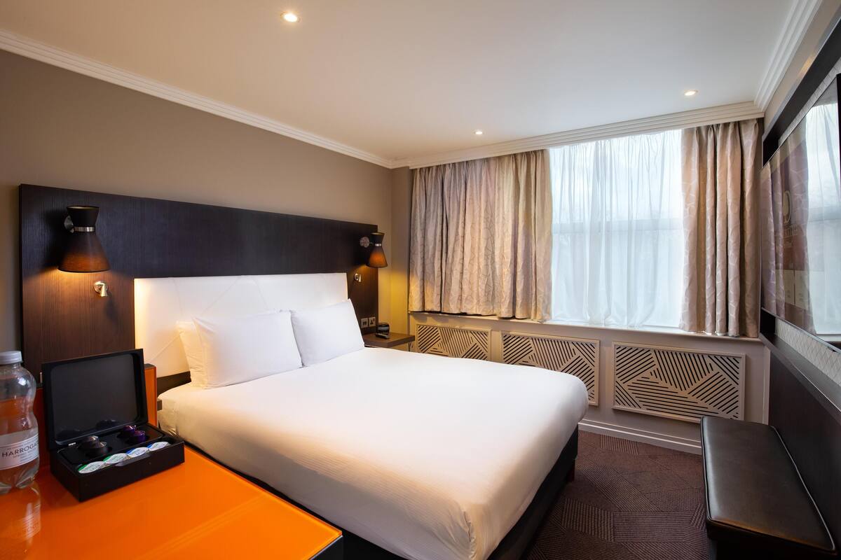 A hotel room with a large bed, a bedside table with a bottle, a small table with a box, a headboard with wall-mounted lights, and a window with curtains.