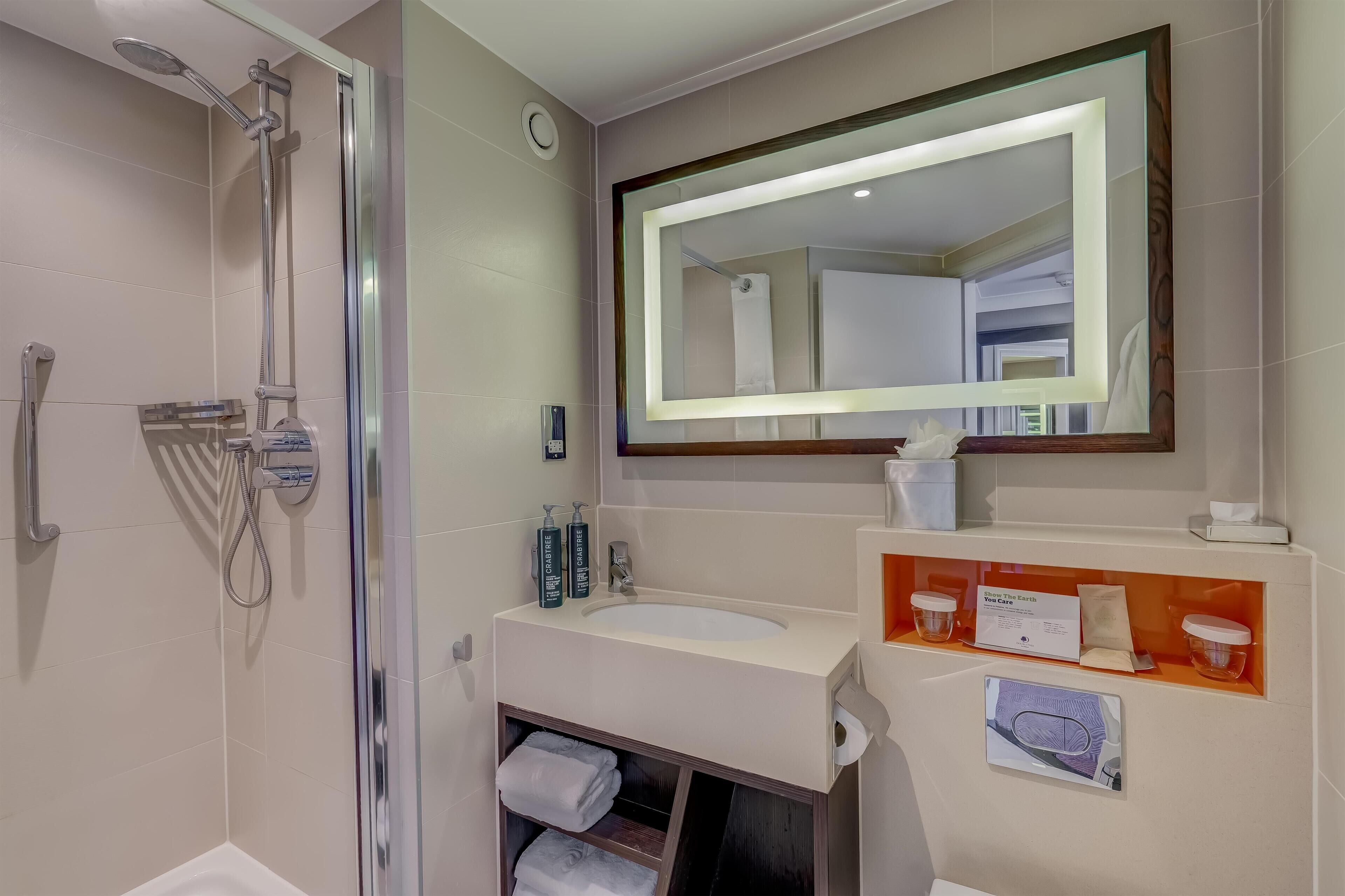 A modern bathroom with a shower, a large mirror, a sink, and a vanity with toiletries.