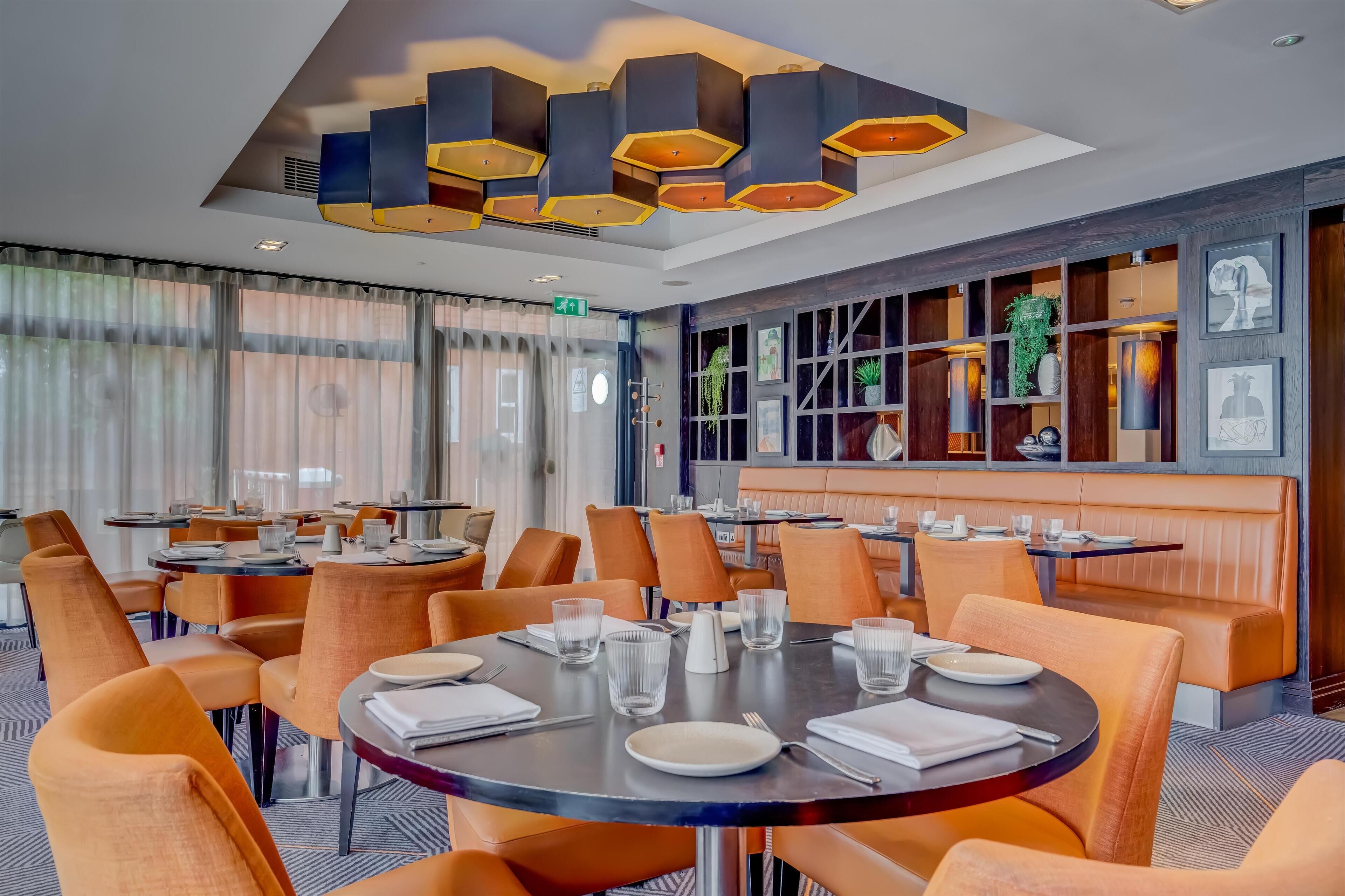 A modern dining area with round tables, orange chairs, and a ceiling with hexagonal light fixtures.