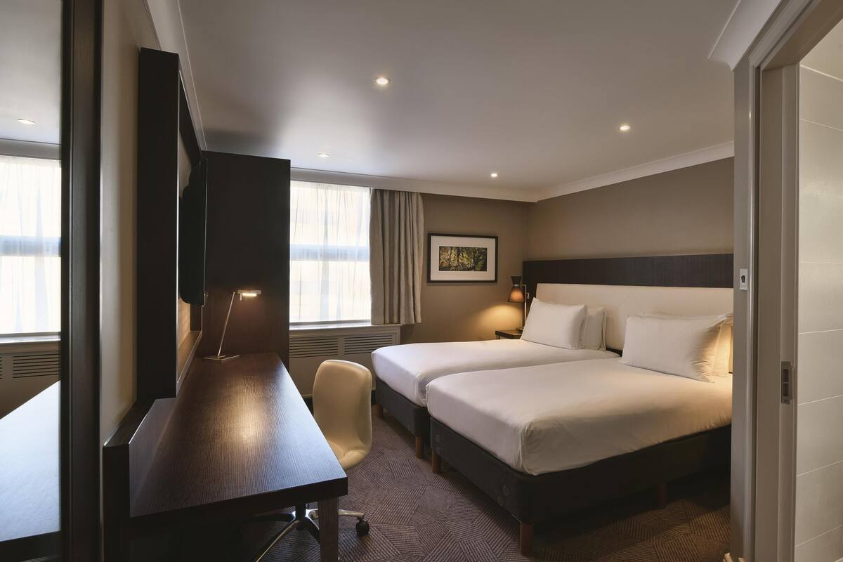 A hotel room with a desk, two beds, a chair, a framed picture on the wall, and a window with curtains.