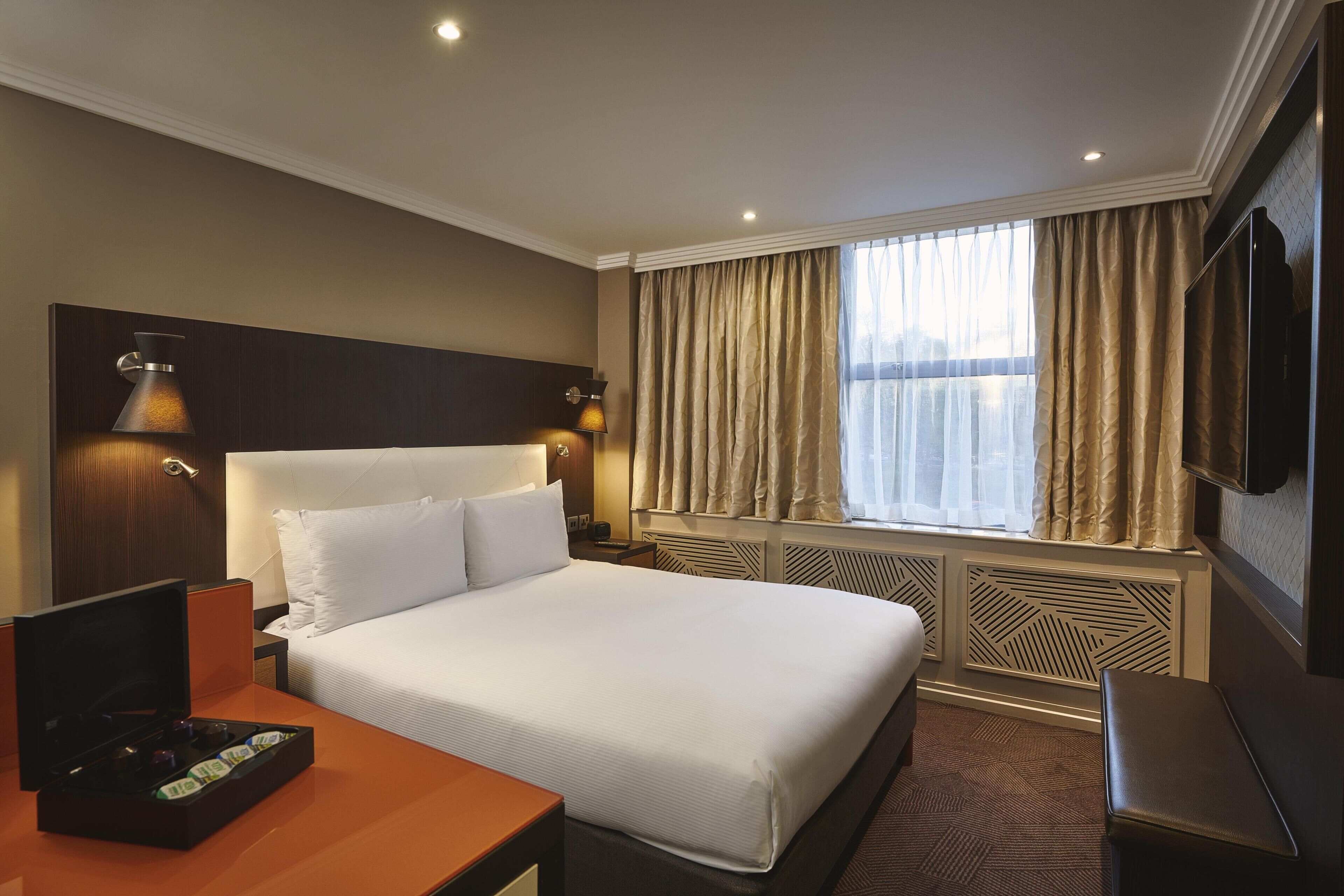 Ein Hotelzimmer mit einem großen Bett, einem Fernseher, einem Schreibtisch mit einem Laptop und einem Fenster mit Vorhängen.