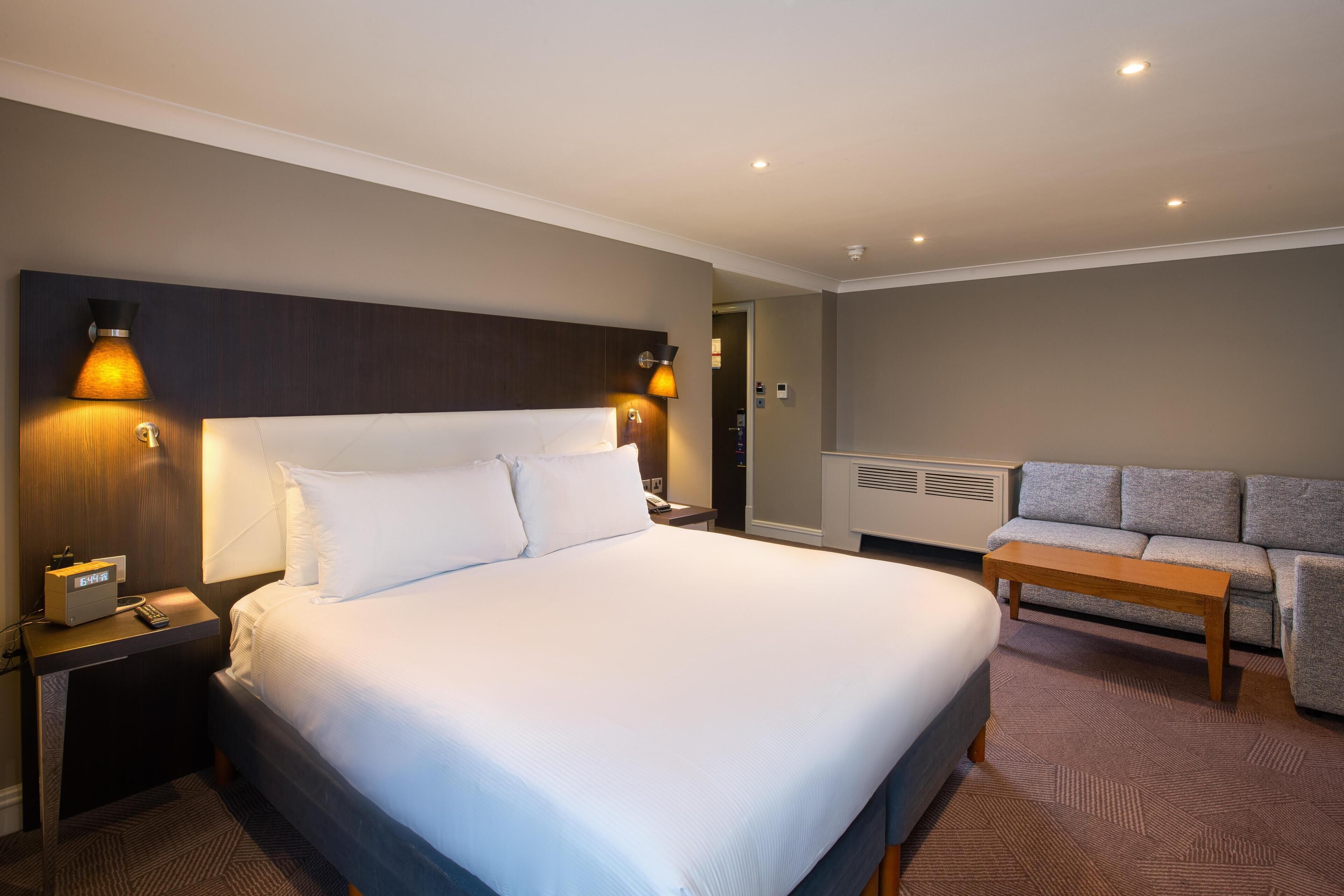 A hotel room with a large bed, bedside lamps, a bedside table with a phone, a sofa, and a small table.