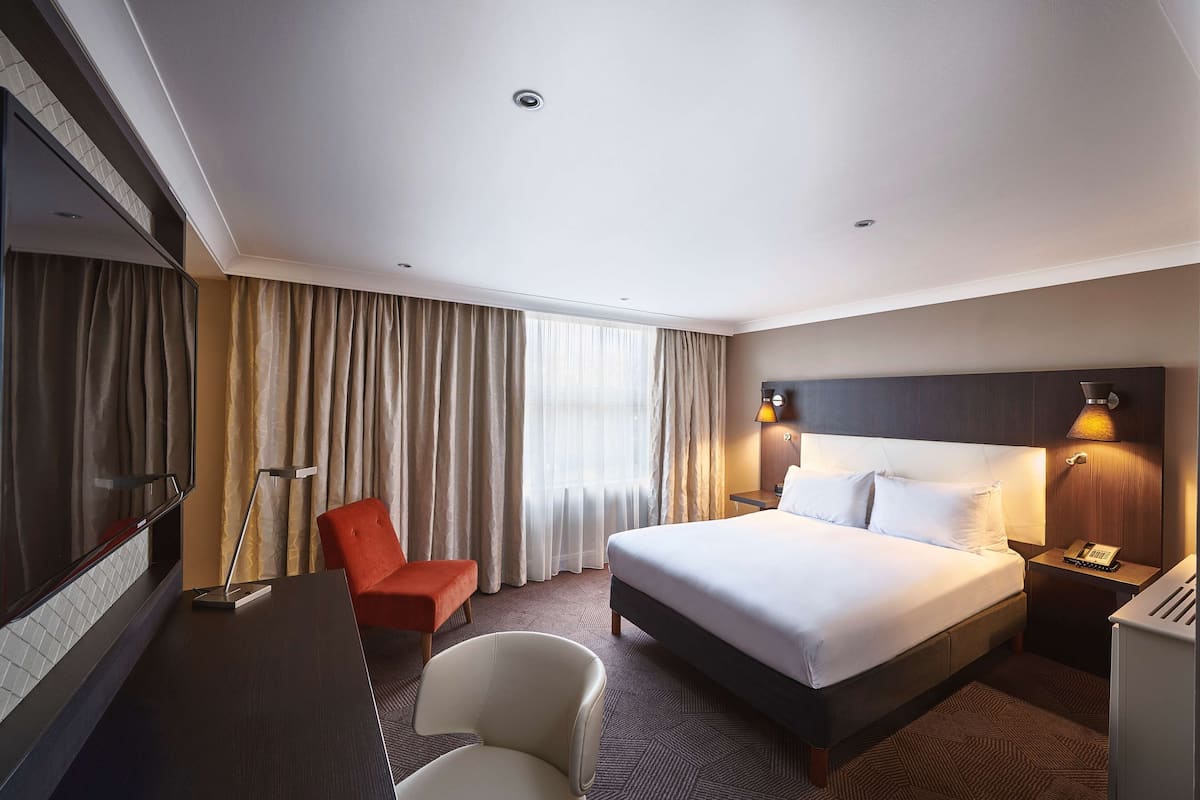 A hotel room with a bed, a desk, a chair, a TV, and a window with curtains.