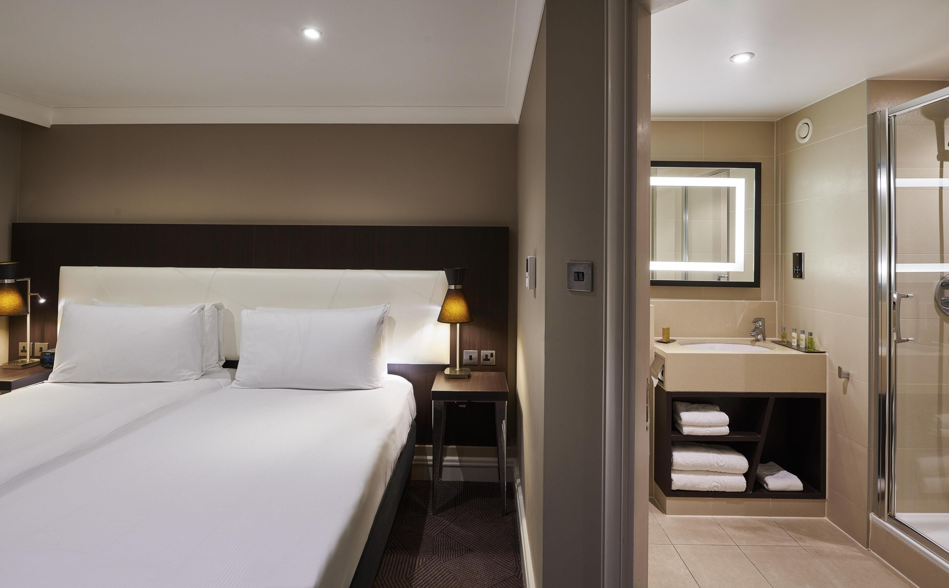 A hotel room with a large bed, bedside lamps, a nightstand, a bathroom with a sink, mirror, and a shower.