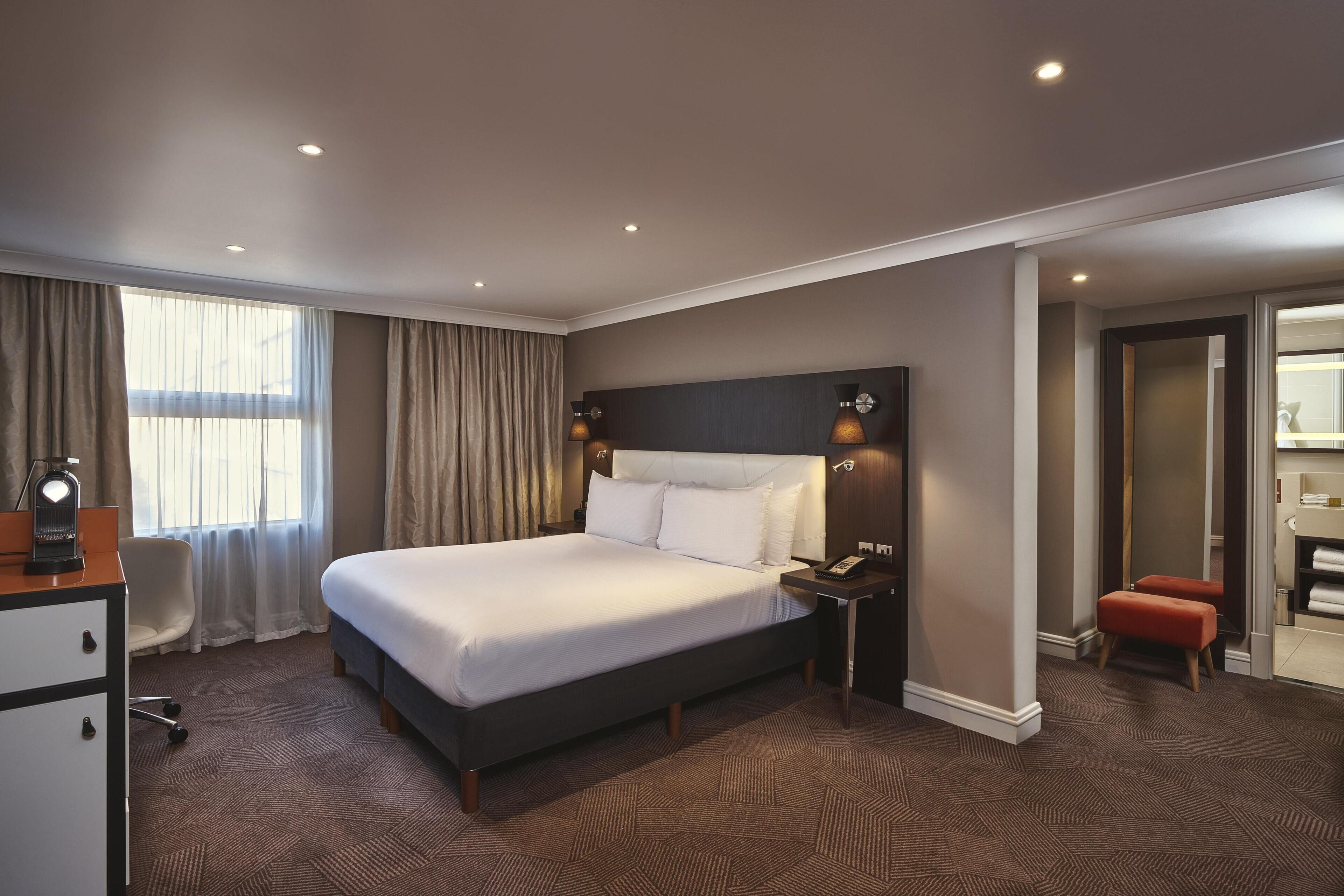 A hotel room with a large bed, a desk, a chair, a nightstand, and a bathroom visible through an open door.