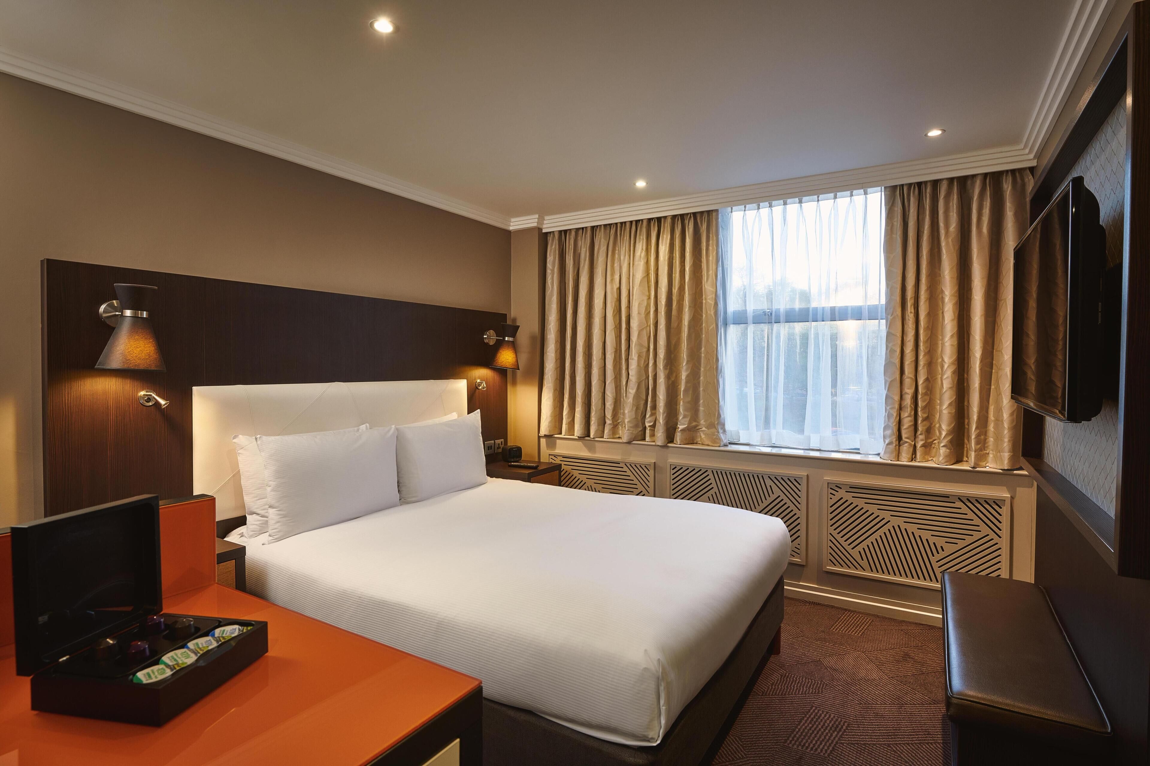 Une chambre d’hôtel avec un grand lit, une télévision, un bureau avec un ordinateur portable et une fenêtre avec des rideaux.